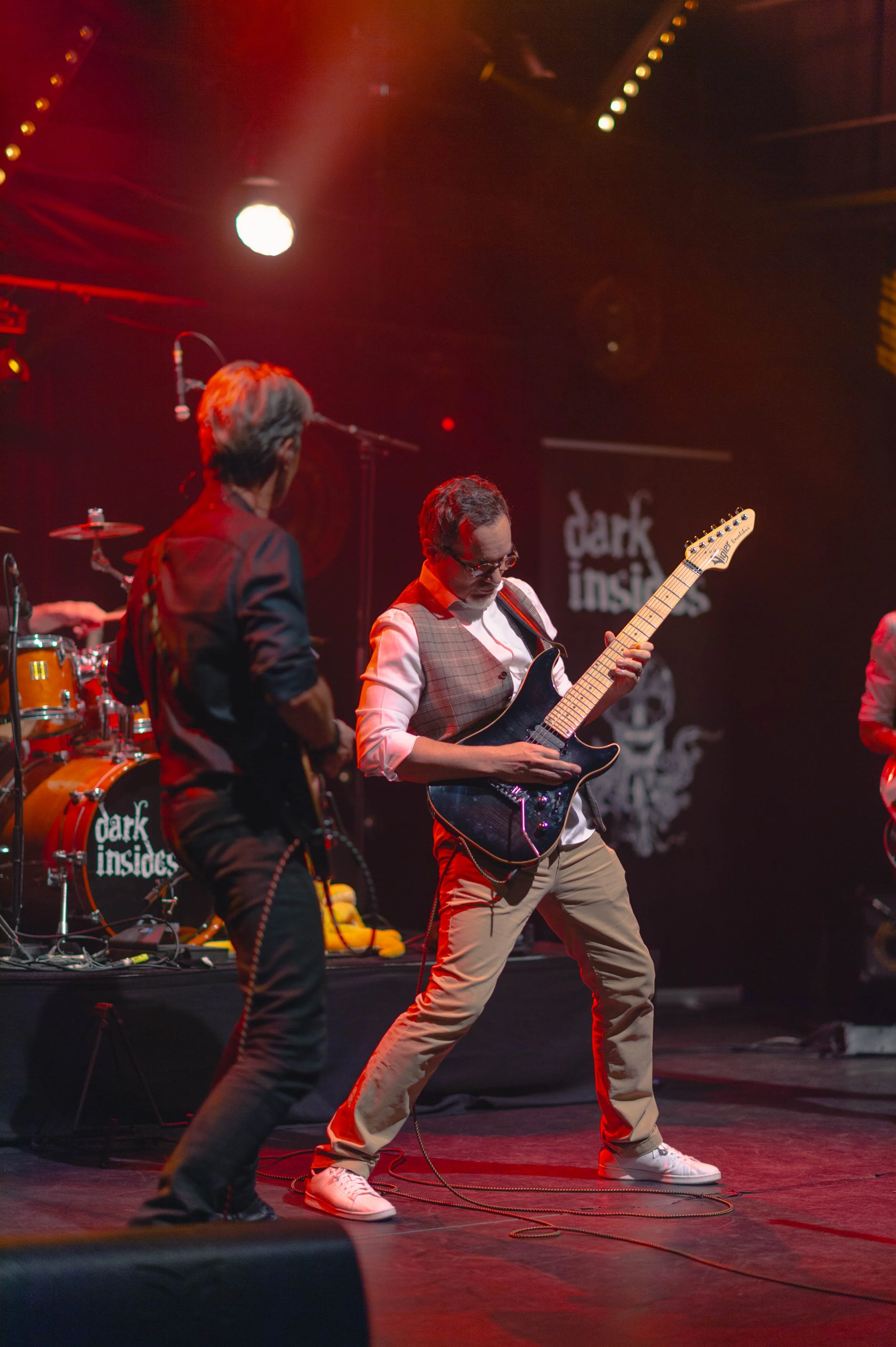 Prise de vue d'un groupe de musiciens en concert sur scène, avec un guitariste principal en avant, portant une veste en gilet et lunettes, en train de jouer de la guitare électrique, avec un arrière-plan lumineux et un logo de groupe 'Dark Inside' su