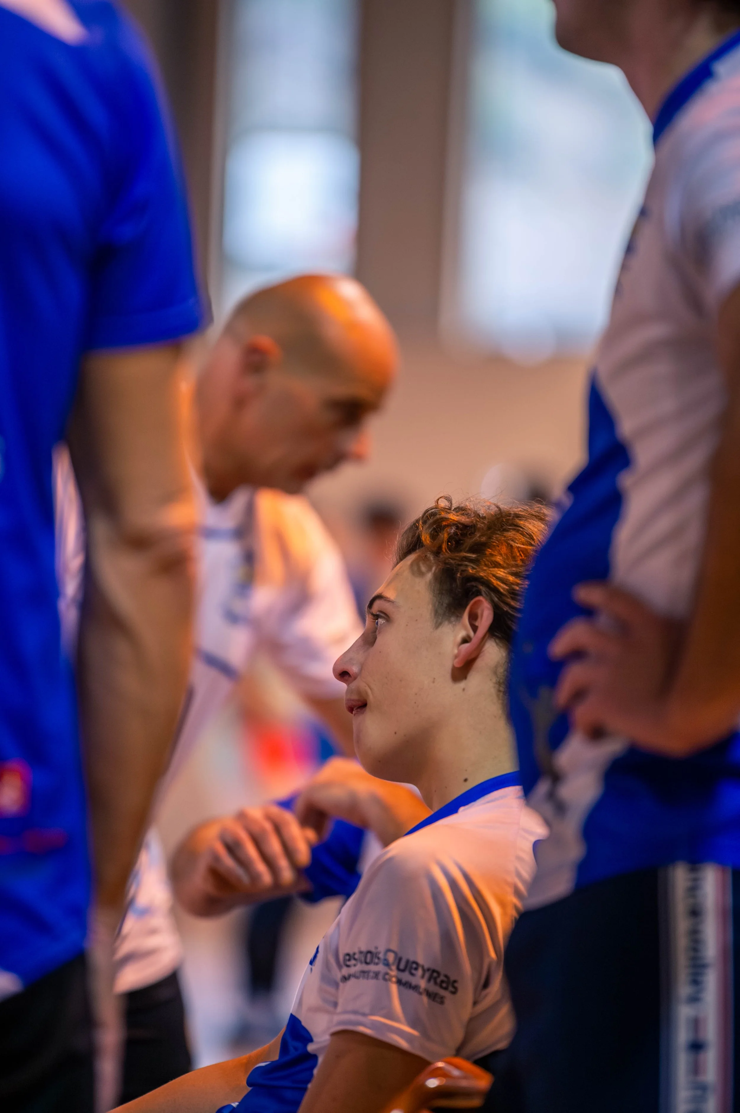 Un jeune homme en maillot de sport blanc et bleu, assis, écoute attentivement des adultes lors d'une pause, probablement lors d'une activité sportive ou d'une compétition.