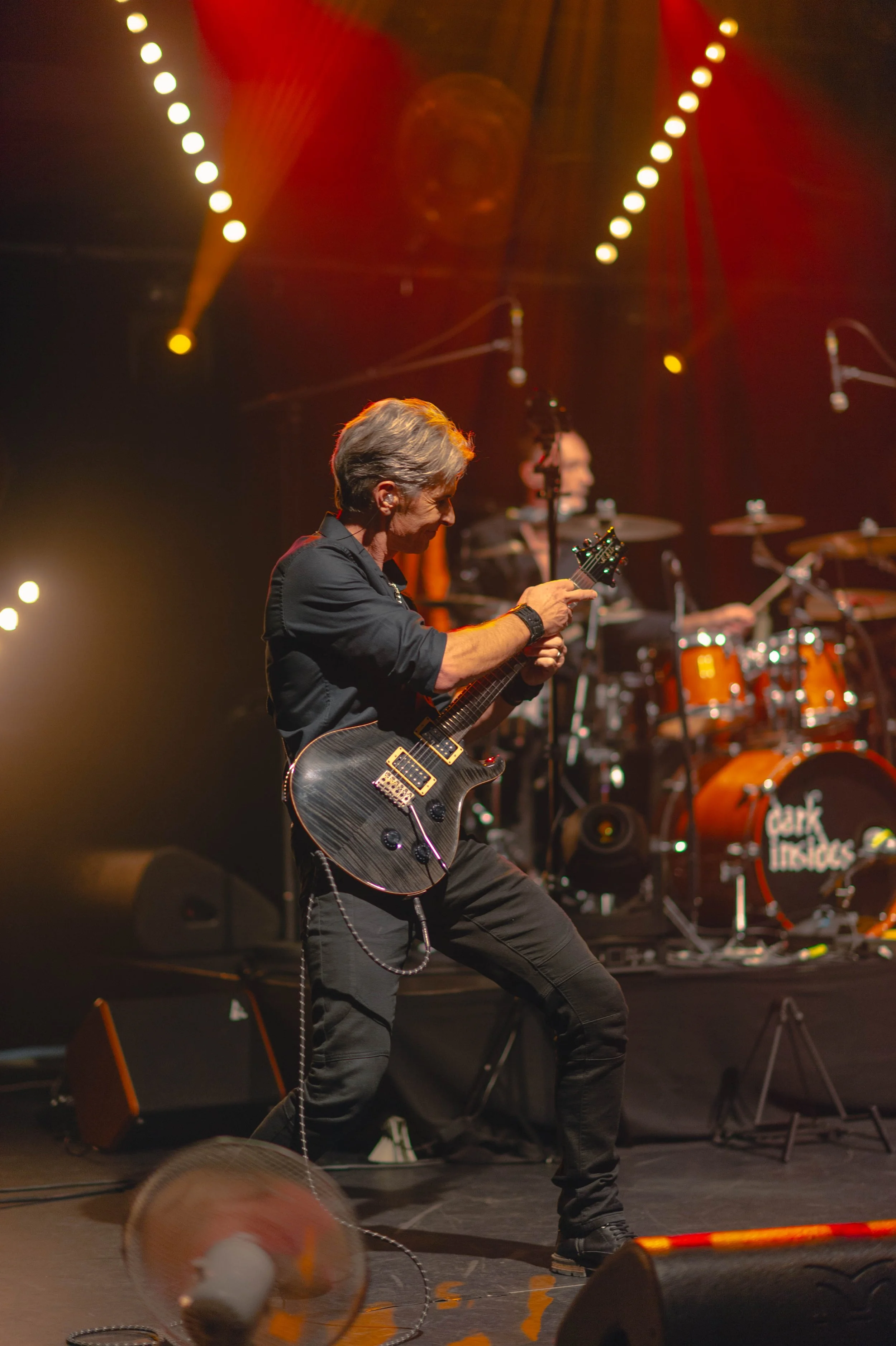 Musicien jouant de la guitare électrique sur scène, avec un batteur en arrière-plan, sous un éclairage de concert.