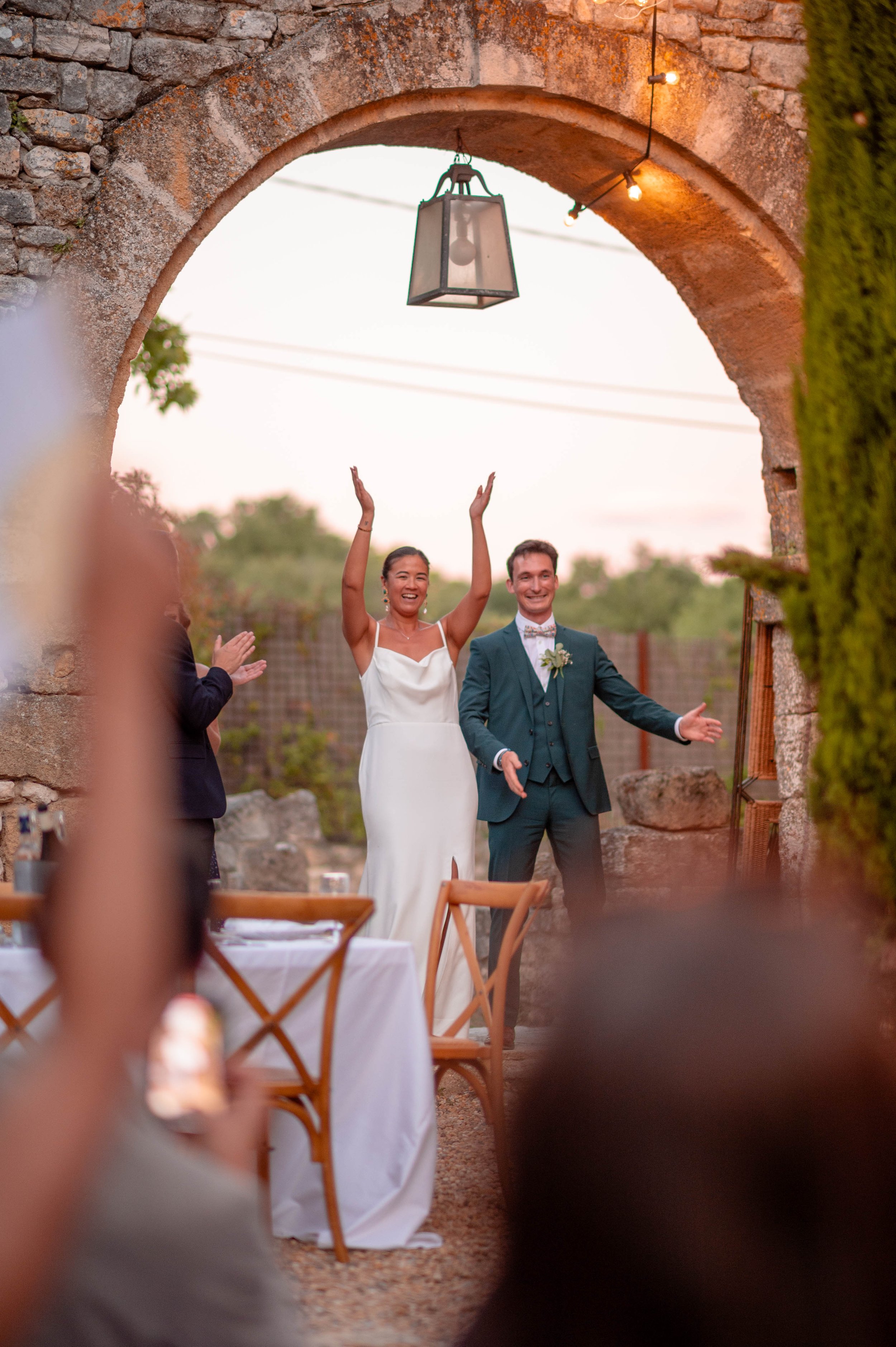 Un couple de mariés célébrant leur mariage sous une arche en pierre à l'extérieur, la mariée en robe blanche avec les bras levés et le sourire, le marié en costume avec un nœud papillon, entourés de convives applaudissant, lors d'une soirée au couche