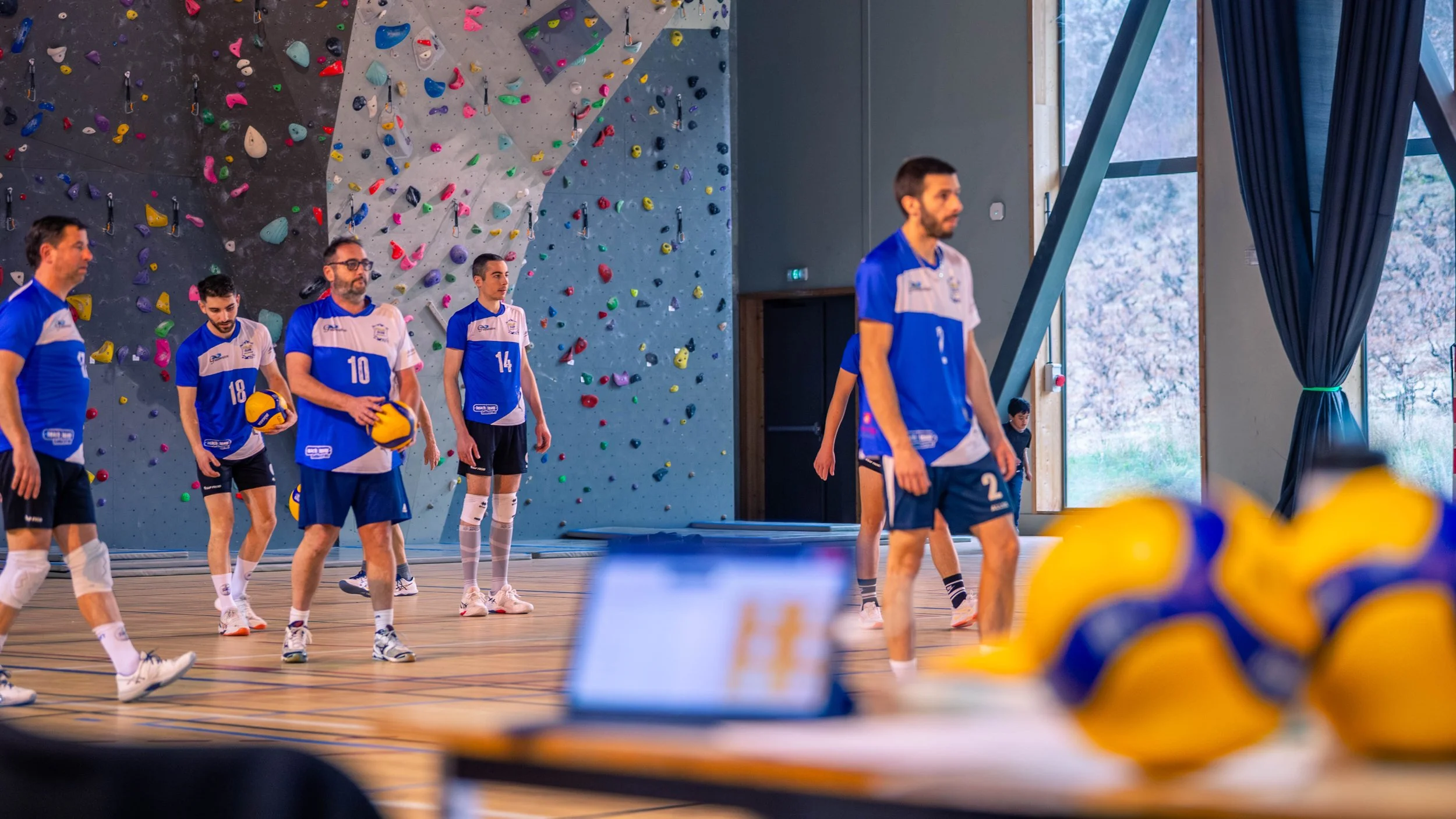 Équipe de volleyball en entraînement dans un gymnase avec mur d'escalade, portant des maillots bleus et blancs, en train de se préparer ou de s'échauffer.