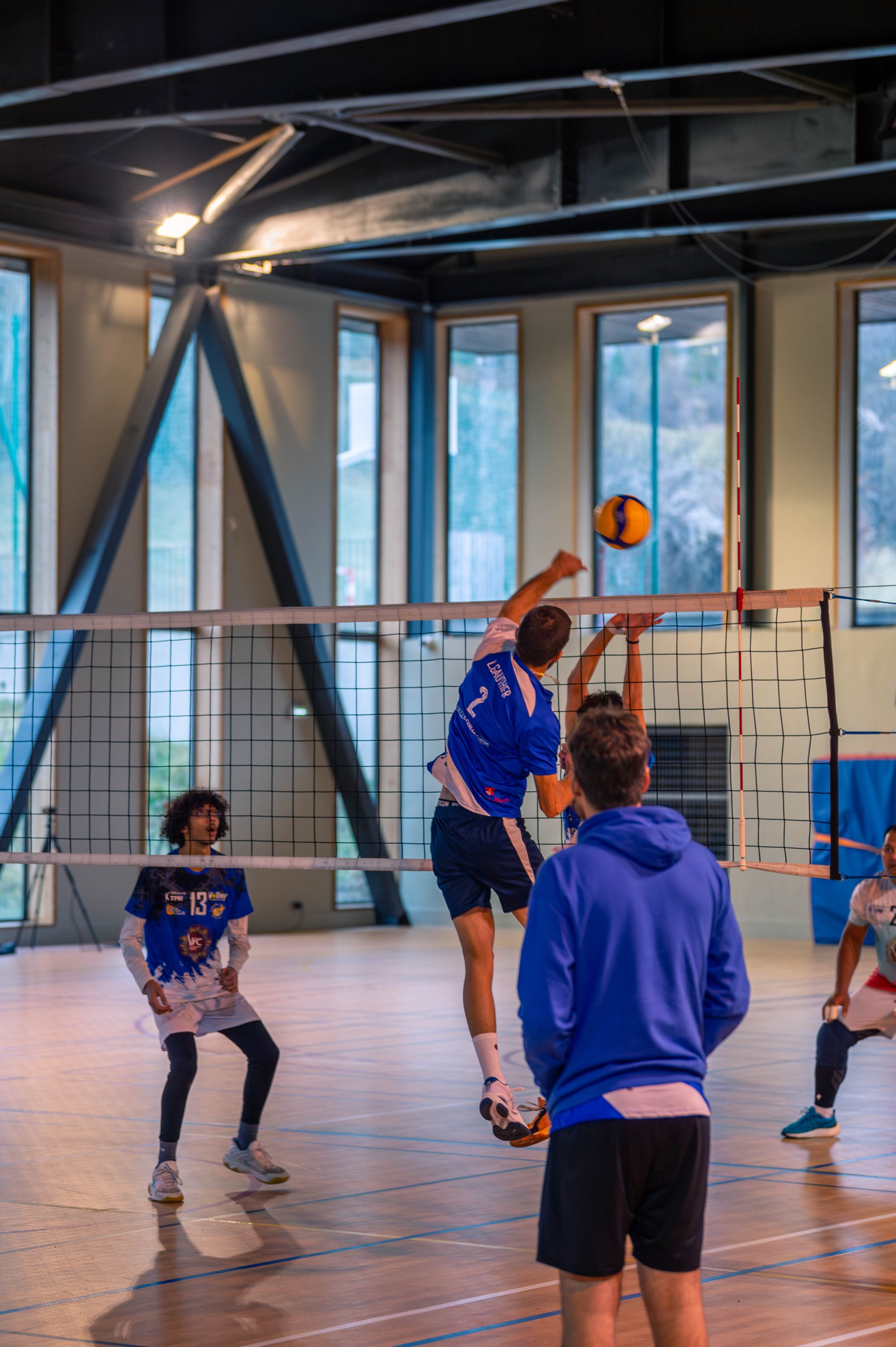 Jeux de volley-ball en intérieur, joueurs en action, un joueur sautant pour frapper le ballon, autres joueurs observant, grands fenêtres en arrière-plan.