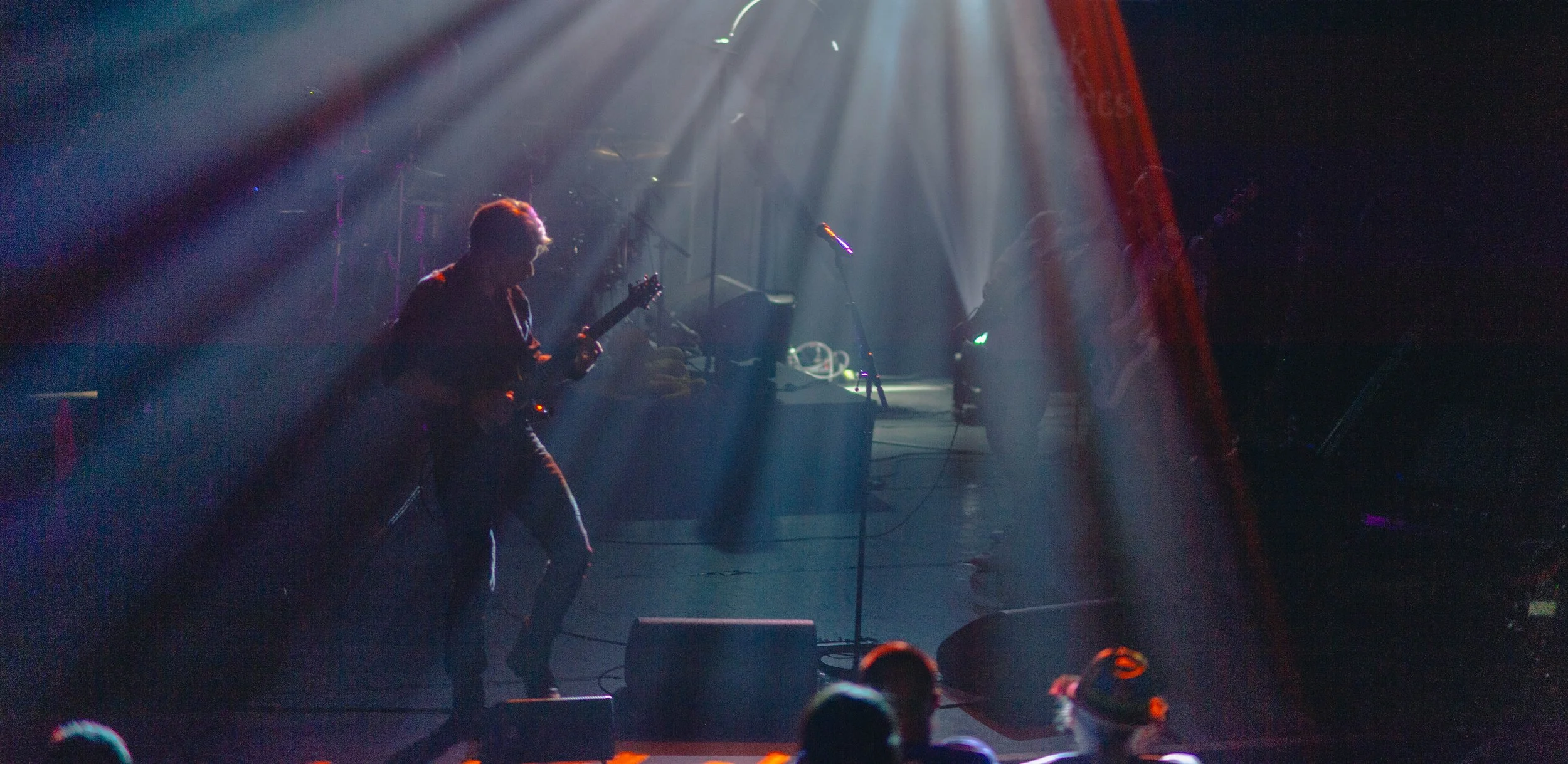 Musiciens jouant sur scène dans une lumière tamisée avec des rayons de lumière visibles projectionant du plafond, ambiance de concert, avec public en premier plan.