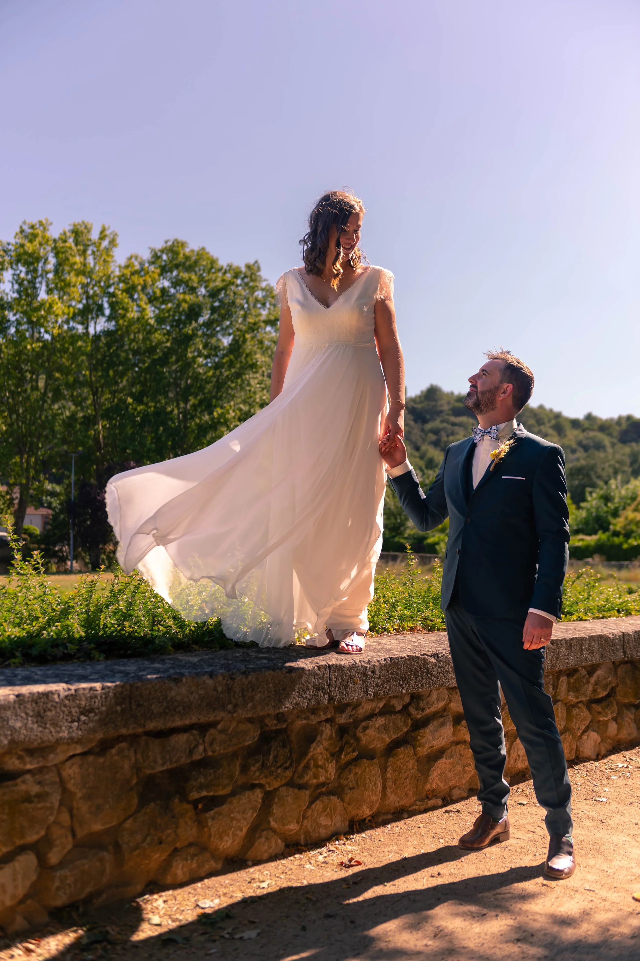 Un couple de mariés lors d'une séance photo en plein air, avec la mariée en robe blanche et le marié en costume noir, la mariée étant sur un mur de pierre et le marié qui l'aide à monter, sous un ciel ensoleillé.
