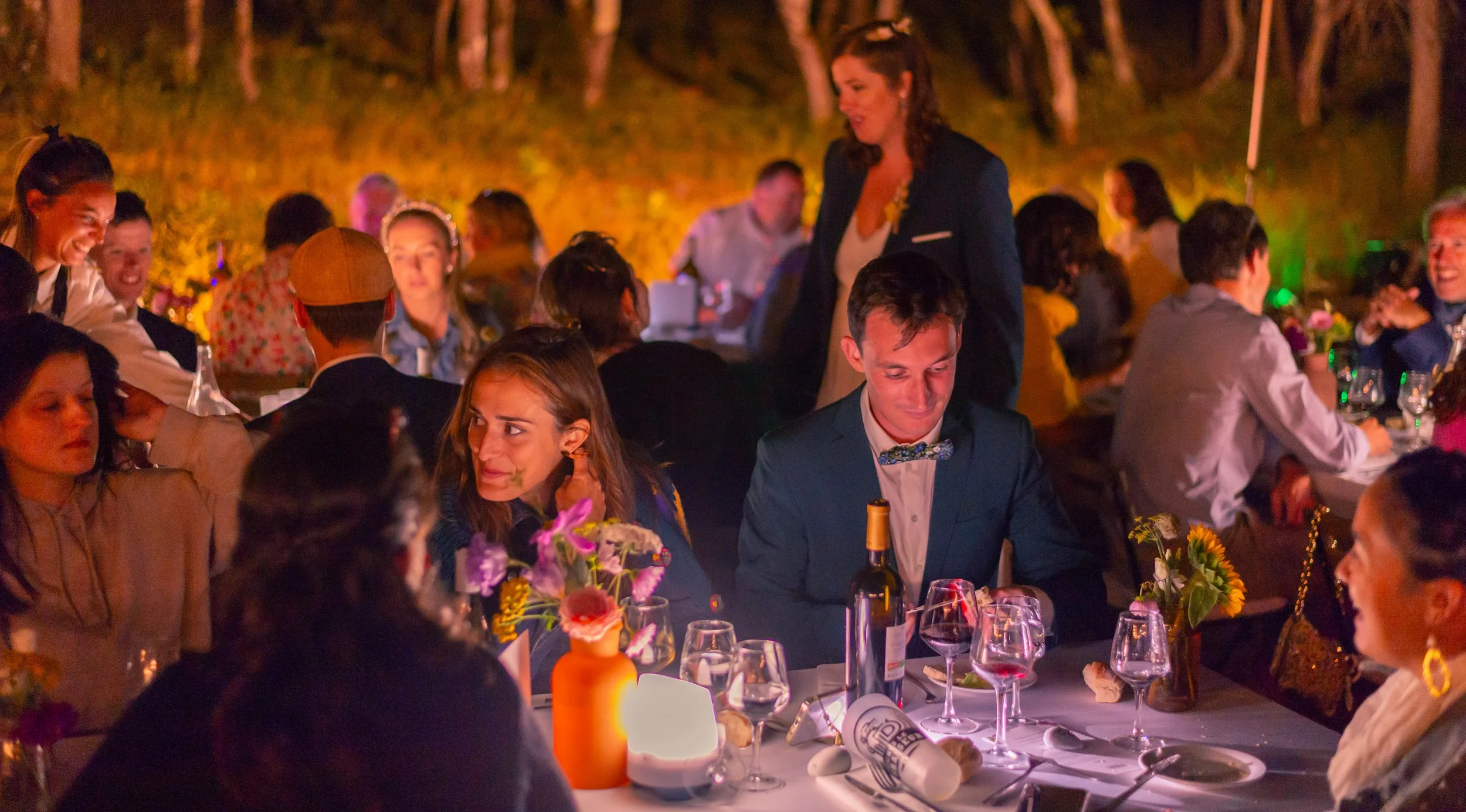 Groupe de personnes assises à une table lors d'une soirée en plein air, avec décorations lumineuses et fleurs, partageant un repas et des moments de convivialité.