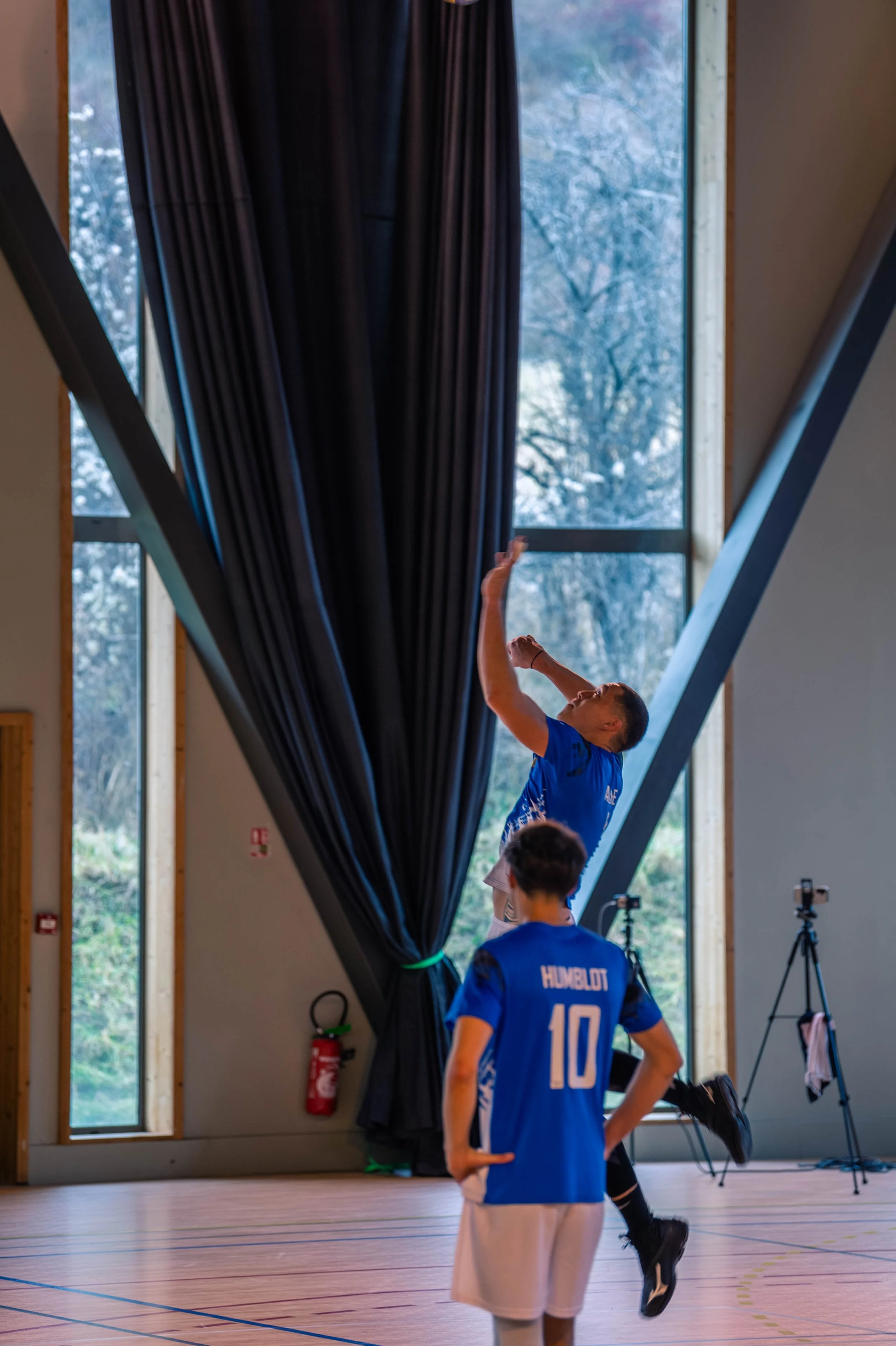 Deux joueurs de handball s'entraînent dans un gymnase, l'un sautant pour lancer et l'autre observant, avec un grand fenêtre derrière eux et un rideau noir, le tout dans un environnement intérieur.