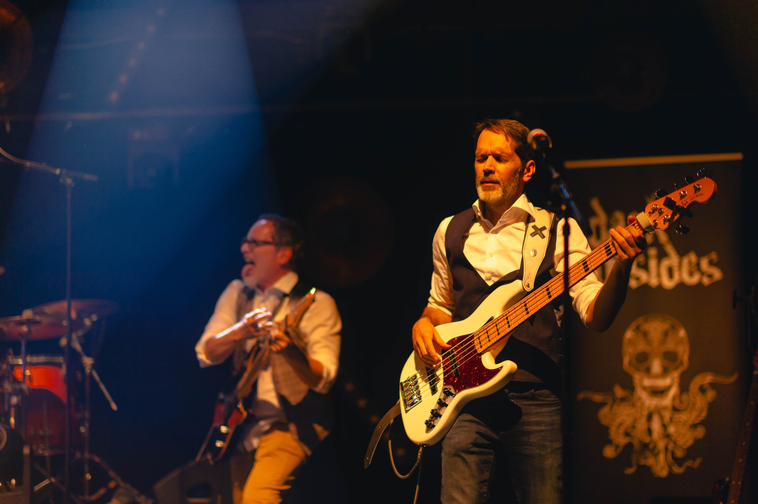 Deux musiciens jouent sur scène, l'un avec une guitare électrique blanche et l'autre avec une guitare acoustique, dans une ambiance sombre avec un éclairage chaud.