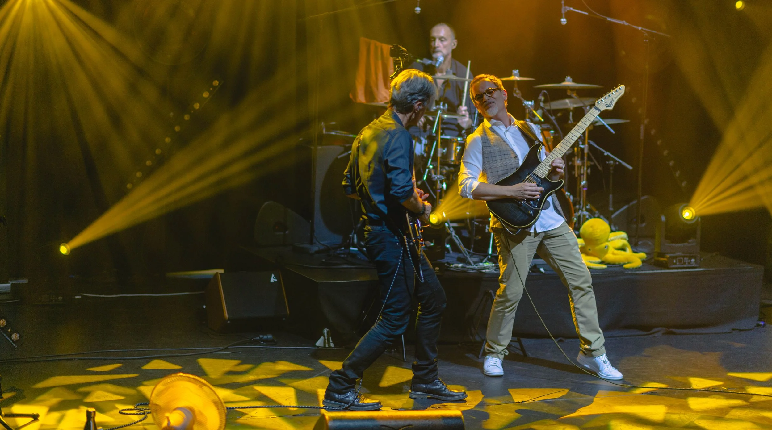 Un groupe de musiciens joue sur scène avec des guitares, un batteur en arrière-plan, et un éclairage jaune dynamique.