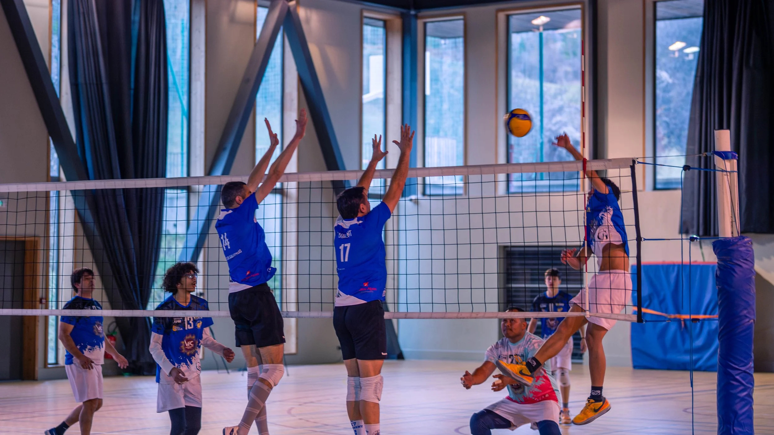 Une partie de volleyball indoor avec des joueurs en action près du filet, dans une salle avec de grandes fenêtres et des rideaux noirs.