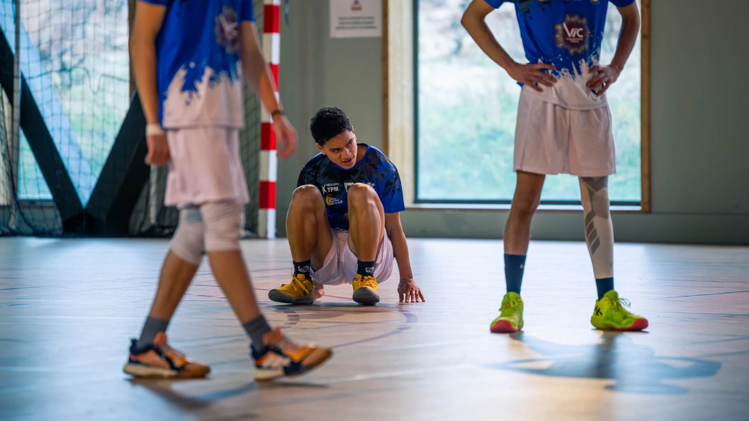 Jeune athlète assis par terre sur un terrain intérieur, entouré d'autres jeunes en tenue sportive, pendant une séance d'entraînement ou un match.