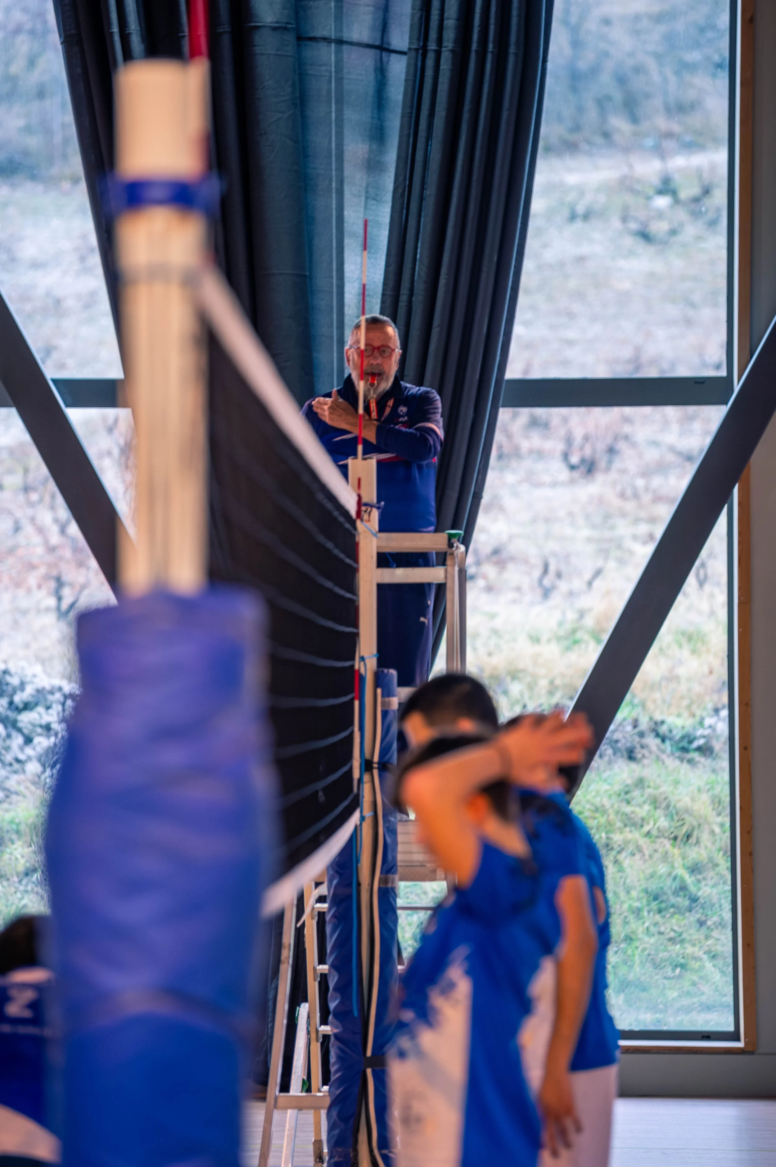 Un entraîneur ou arbitre en tenue entraîne des joueurs de volleyball dans une salle intérieure, avec un filet de volleyball visible. La scène se déroule près d'une grande fenêtre naturelle.