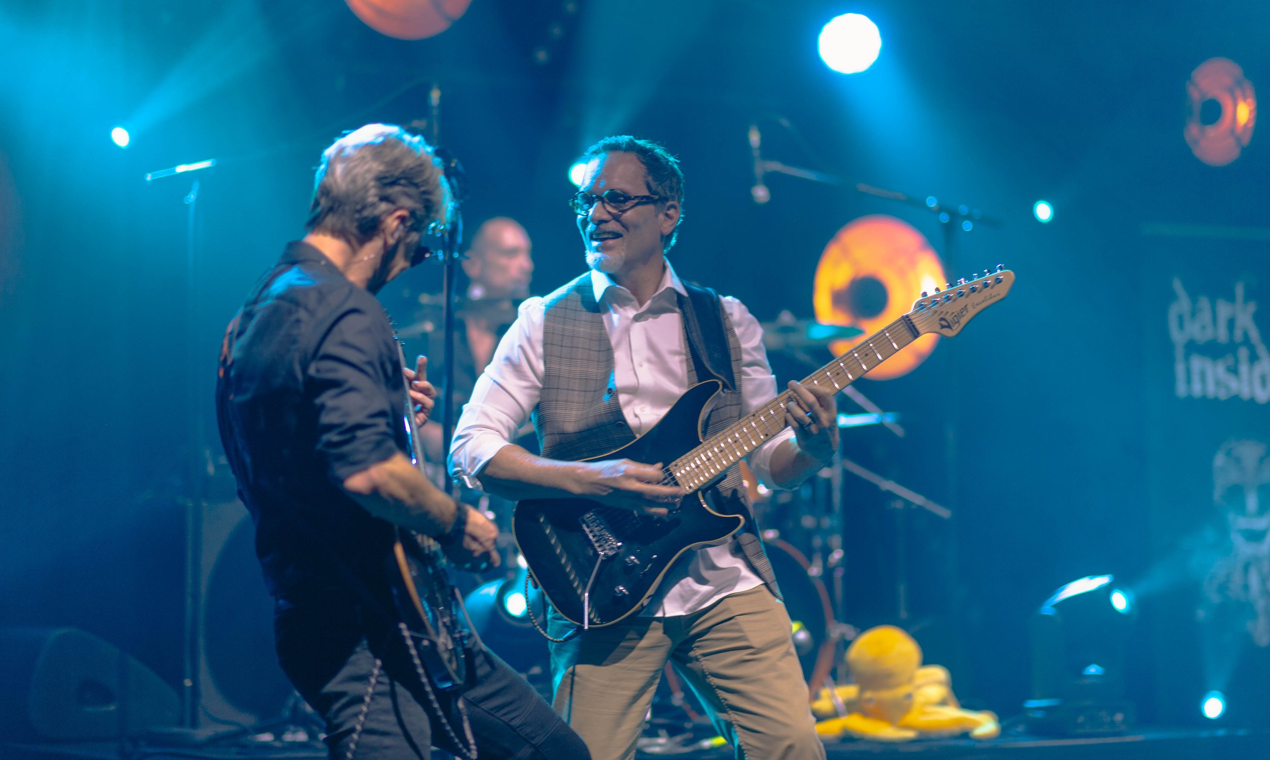 Deux musiciens en concert, un joue de la guitare électrique, l'autre semble chanter ou jouer d'un instrument. Éclairage coloré, arrière-plan avec des lumières et des objets décoratifs.