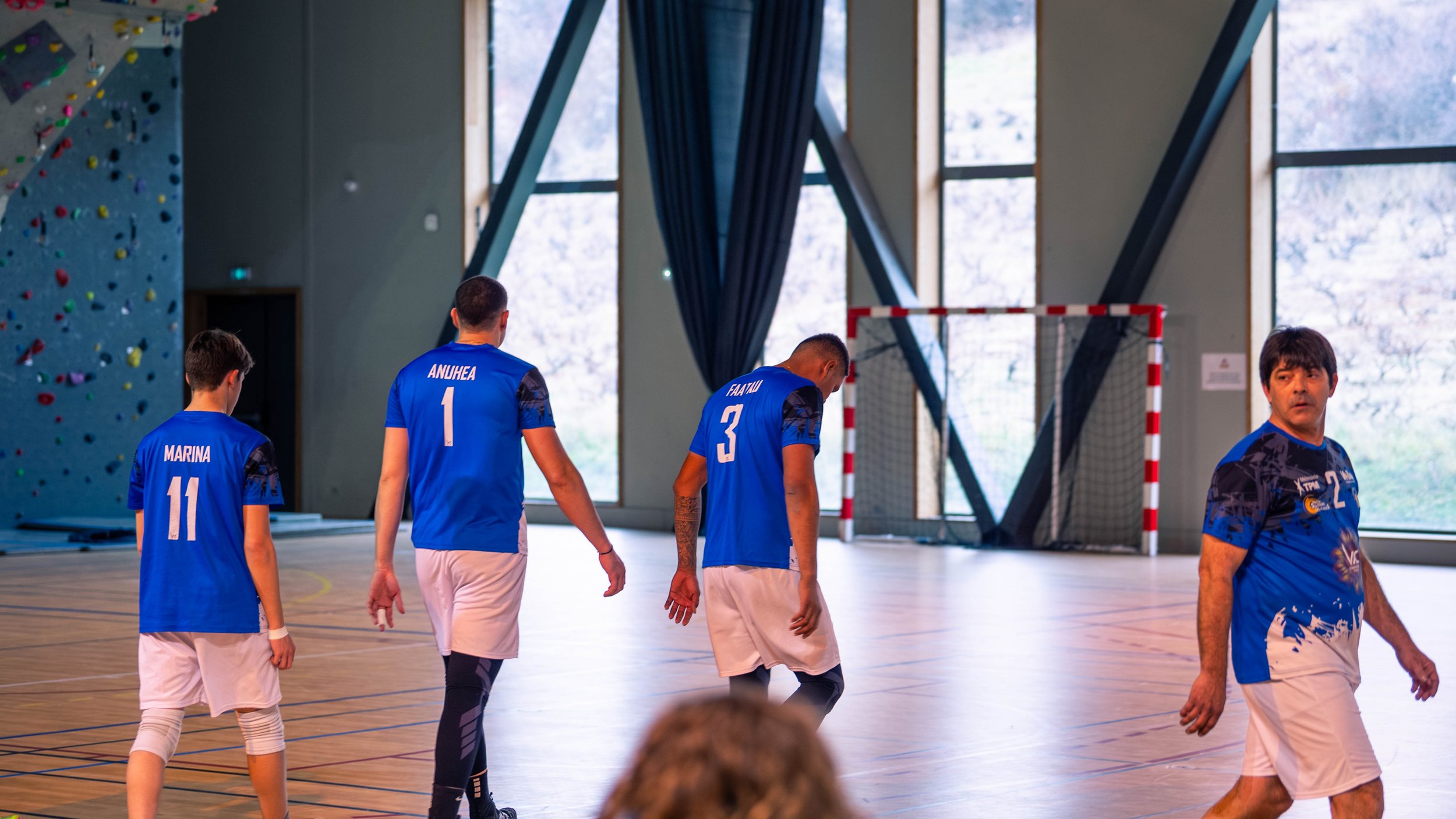 Groupe de joueurs de handball en entraînement ou match dans une salle avec grande fenêtre et terrain en bois.