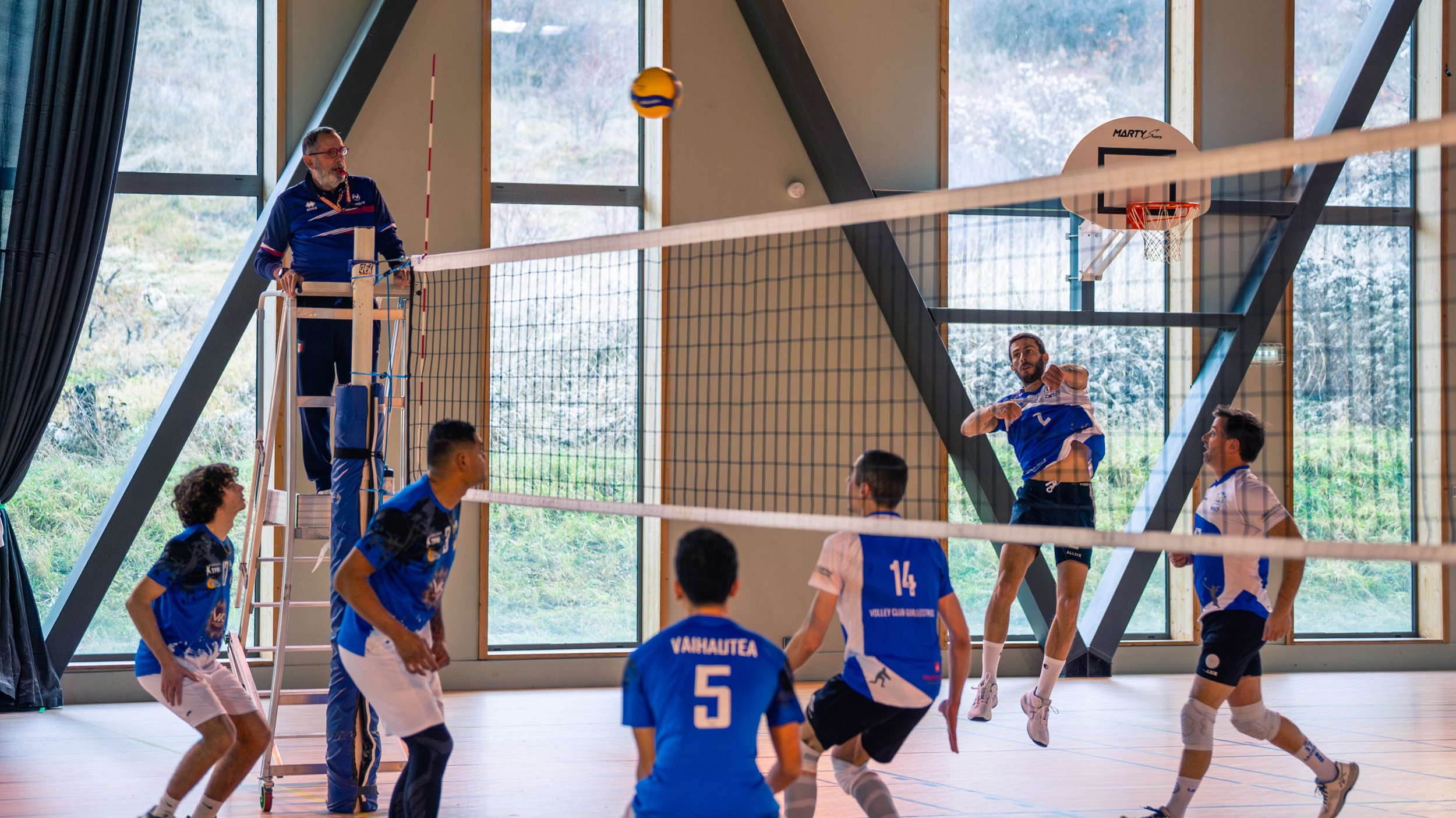 Une équipe de volley-ball en action à l'intérieur d'une salle de sport avec un filet, un arbitre en haut, et des fenêtres en arrière-plan, l'équipe porte des maillots bleus et blancs.