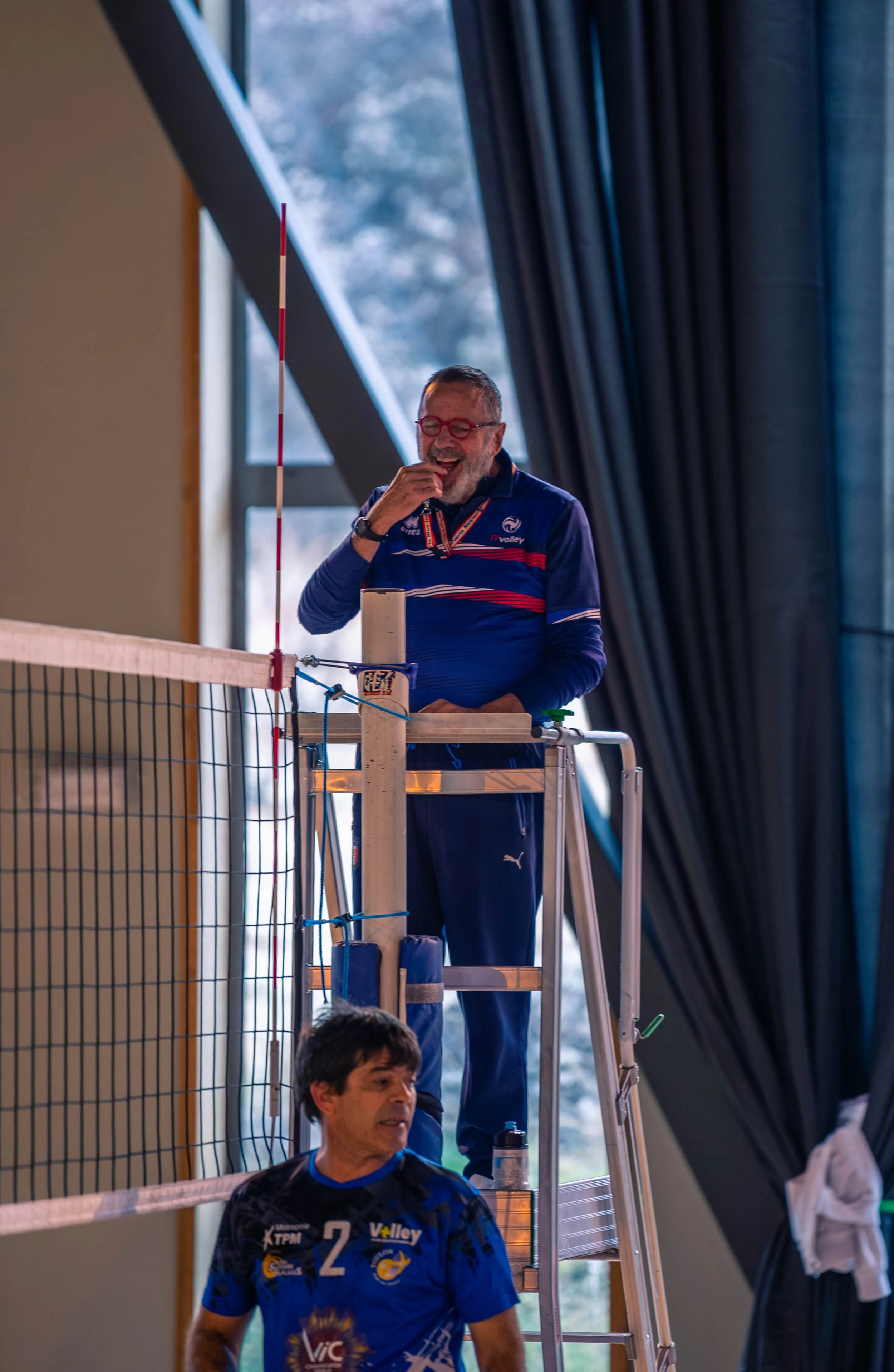 Deux hommes portant des tenues de volleyball, l'un debout sur une plateforme en hauteur mangant quelque chose, l'autre en bas, regardant vers l'avant, dans une salle avec des rideaux et une grande fenêtre.