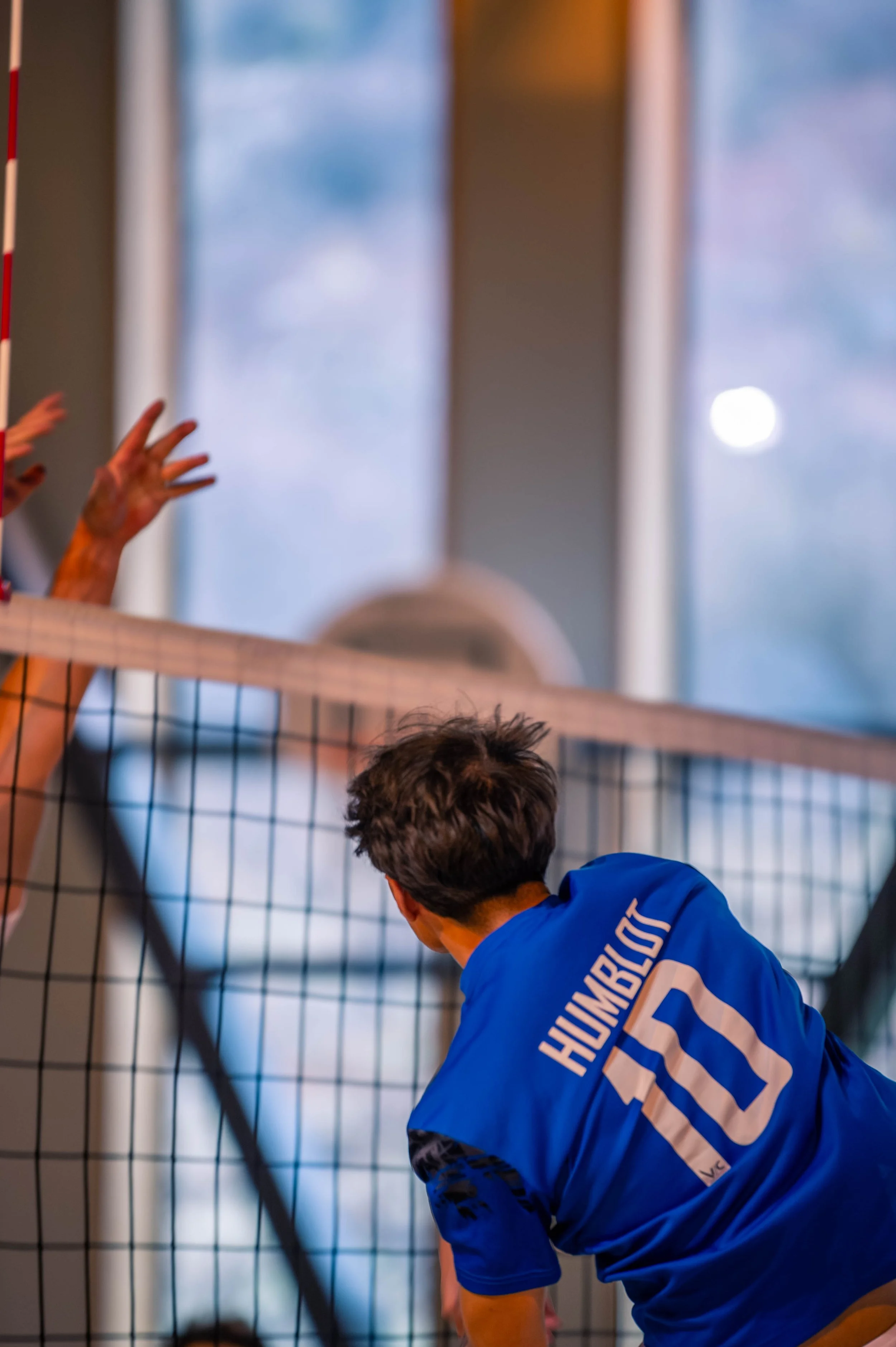 Un joueur de volleyball portant un maillot bleu avec le nom 'HUMBERG' et le numéro 10, en train de faire une action lors d'un match en intérieur, avec une équipe adverse visible de l'autre côté du filet.