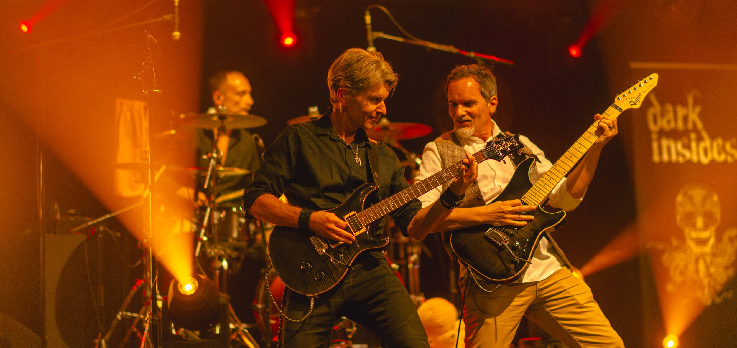 Deux musiciens jouent de la guitare électrique sur scène lors d'un concert, avec un batteur en arrière-plan.