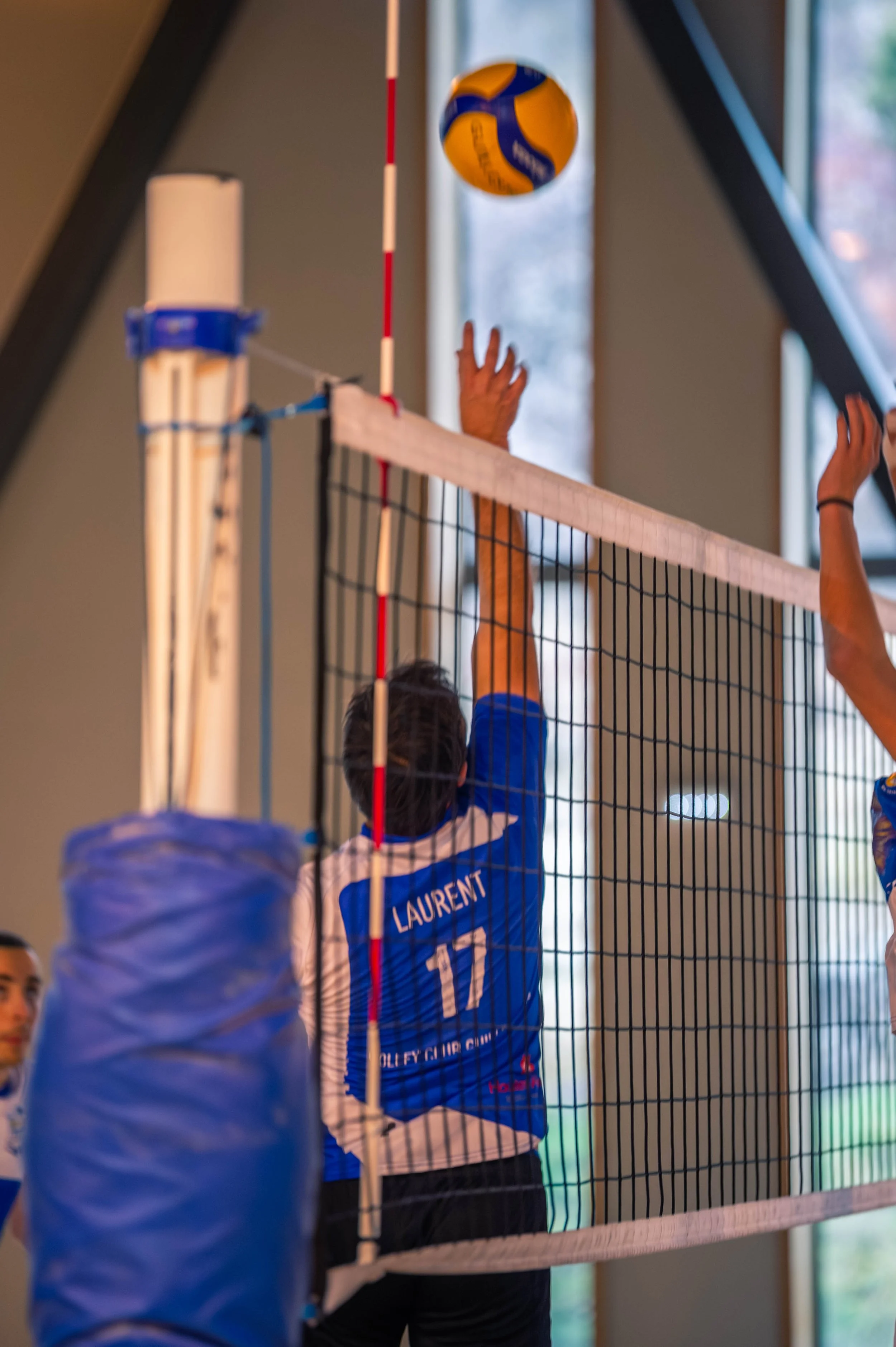 Un joueur de volley-ball portant un maillot avec le nom 'Laurent' et le numéro 17 saute pour contrer une balle au-dessus du filet, dans une salle intérieure.