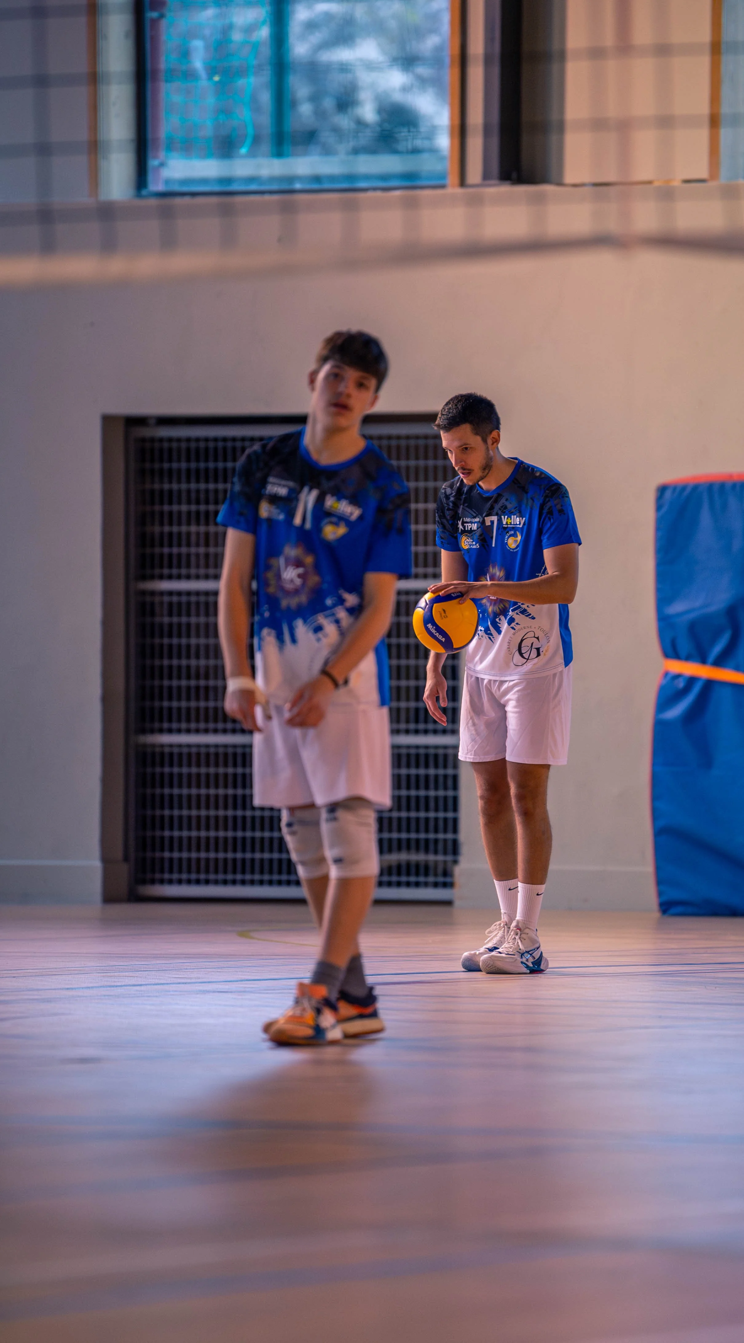 Deux jeunes hommes dans une salle de sport ou gymnase, portant des tenues de volleyball, l'un tenant un ballon, l'autre en arrière-plan, prêts à jouer.
