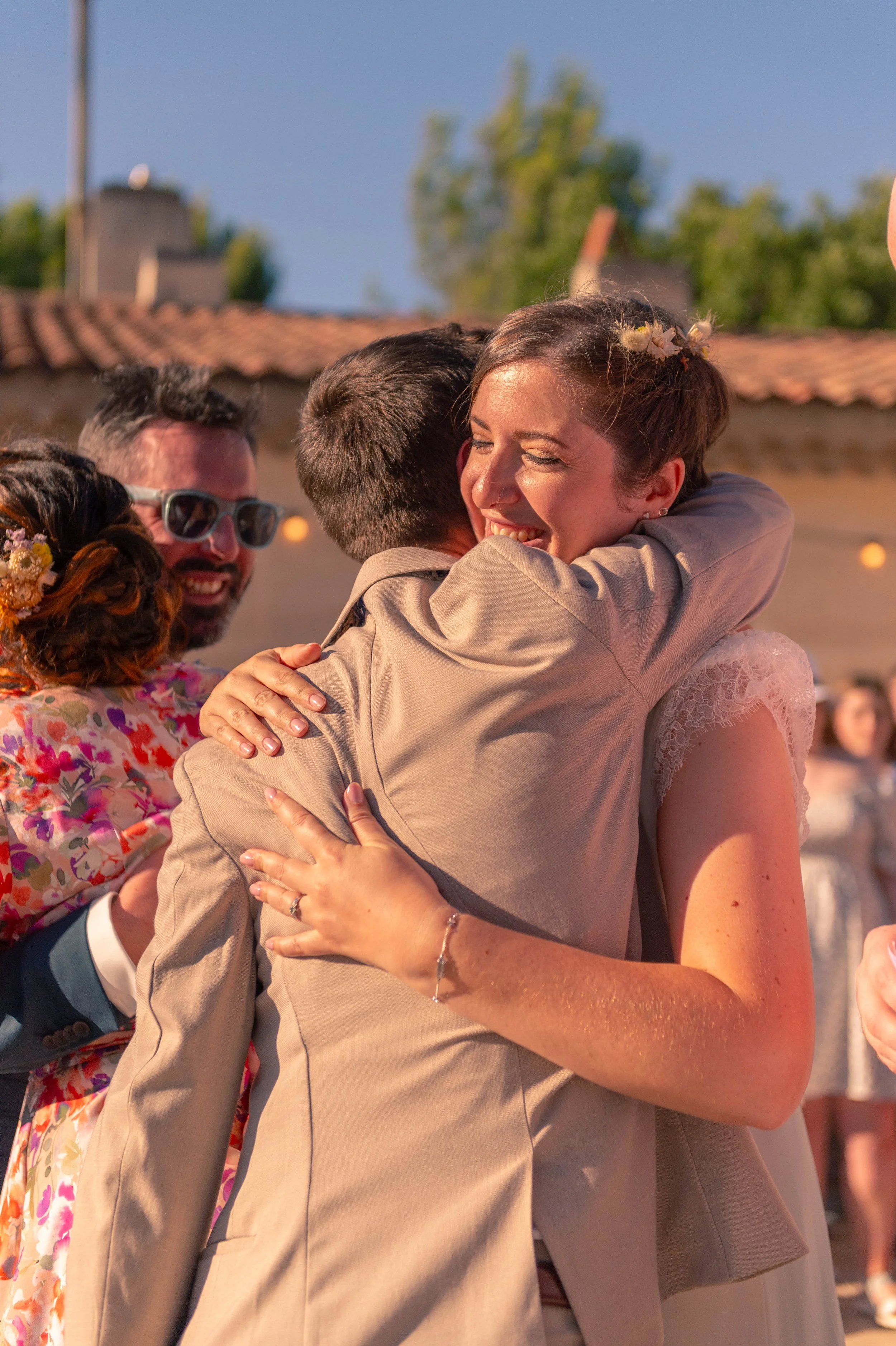 Un couple s'embrasse lors d'une célébration de mariage, entouré de proches, sous un ciel bleu avec un bâtiment en arrière-plan.