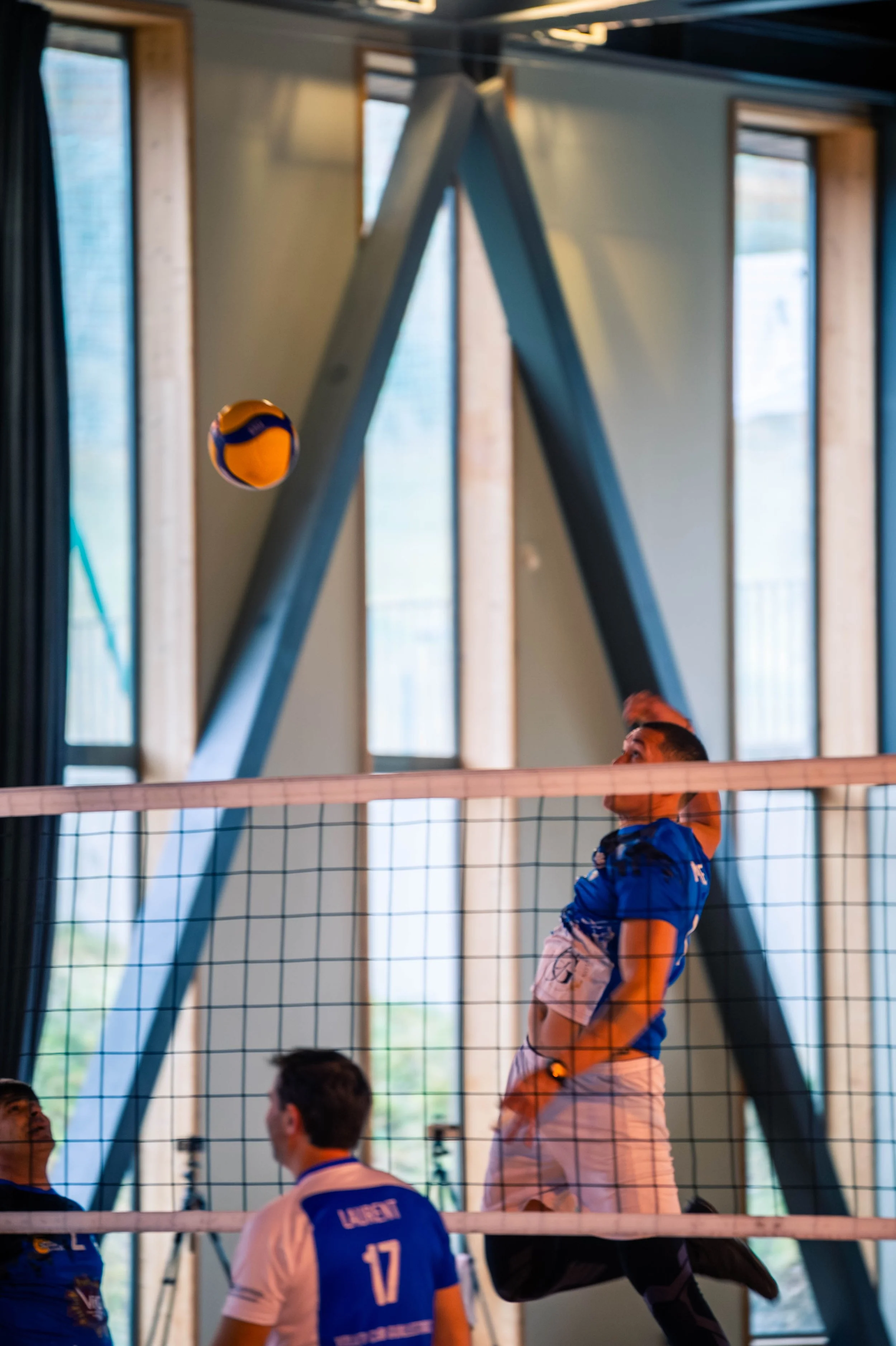 Joueur de volley-ball en vol au-dessus du filet, tentant de frapper le ballon, avec deux autres joueurs en attente en dessous dans un gymnase aux grandes fenêtres.