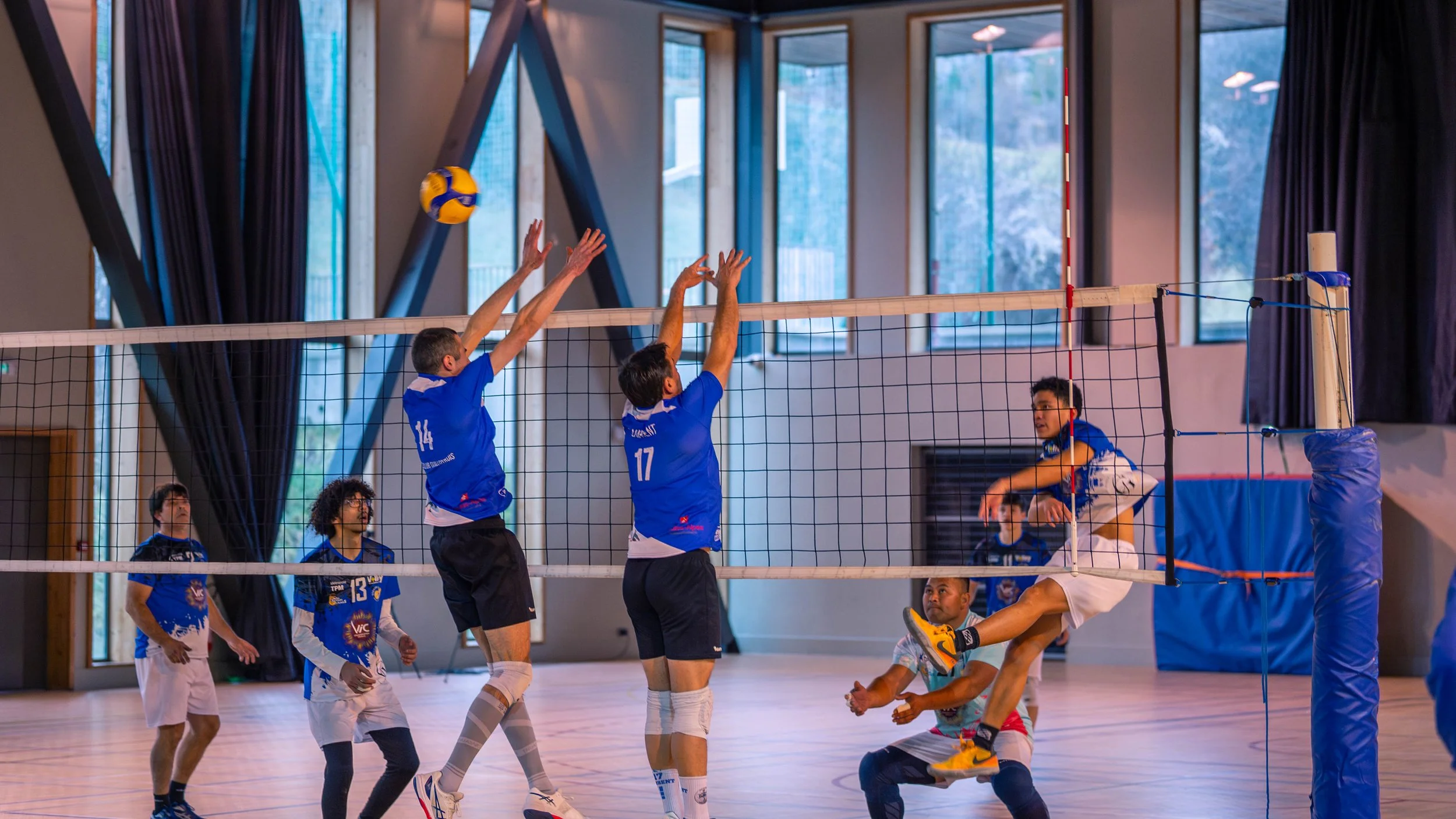 Des joueurs de volleyball en action à l'intérieur d'une salle, avec un joueur en train de sauter pour attaquer ou bloquer, tandis que d'autres tentent de contrer ou de défendre, le tout sous une lumière tamisée.