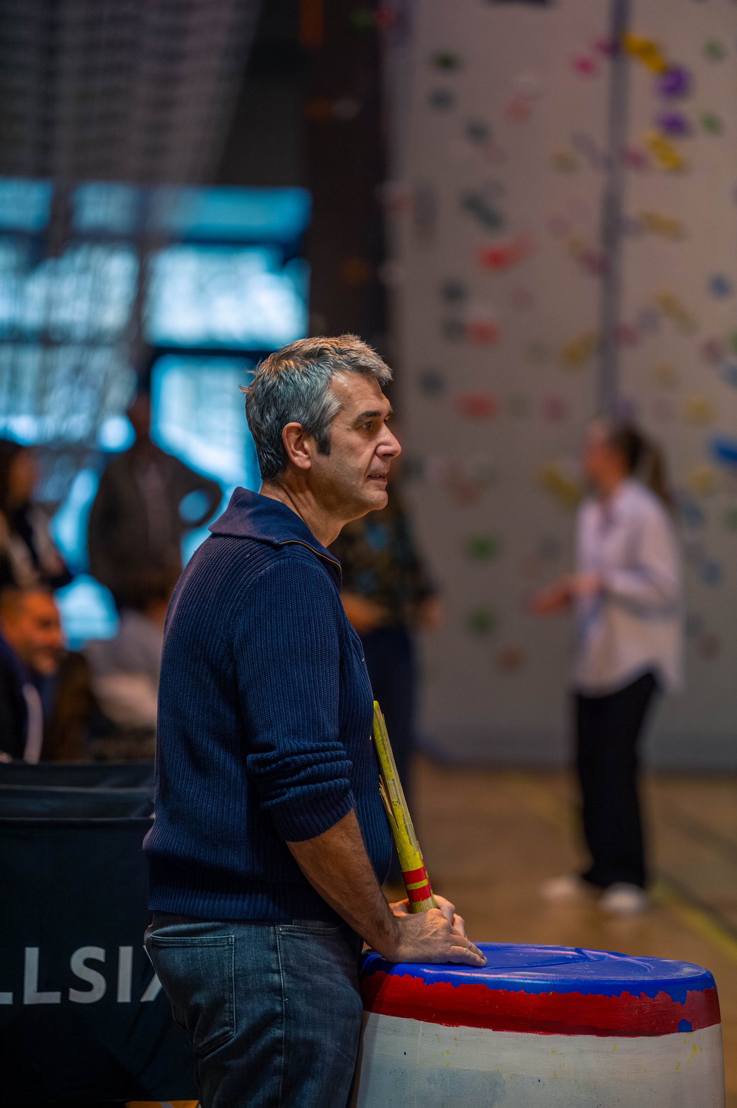 Un homme d'âge moyen en pull bleu regarde dans une salle d'escalade où des personnes s'entraînent.