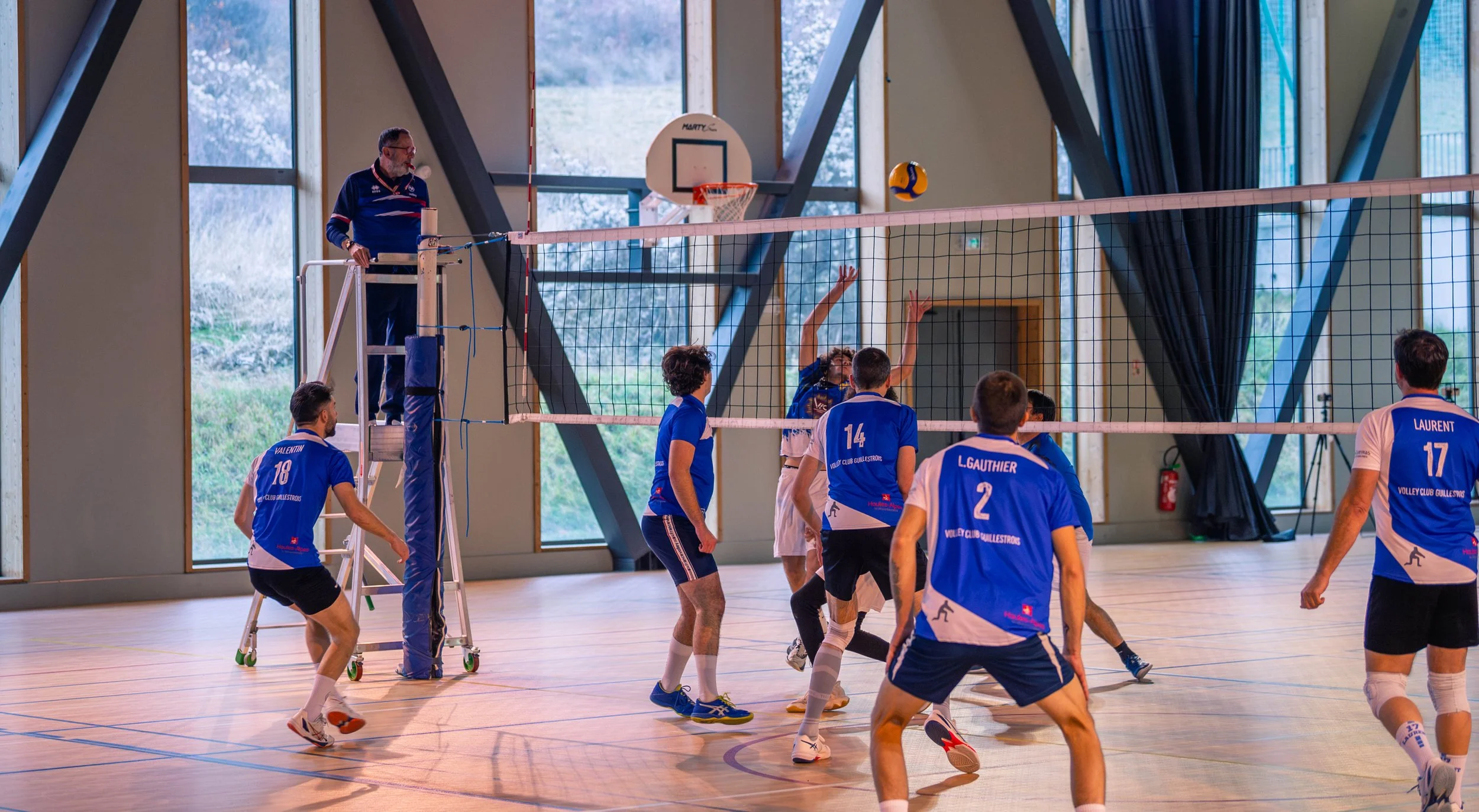Une equipe de volleyball jouant dans un gymnase intérieur avec un arbitre sur une plateforme.et une balle volante