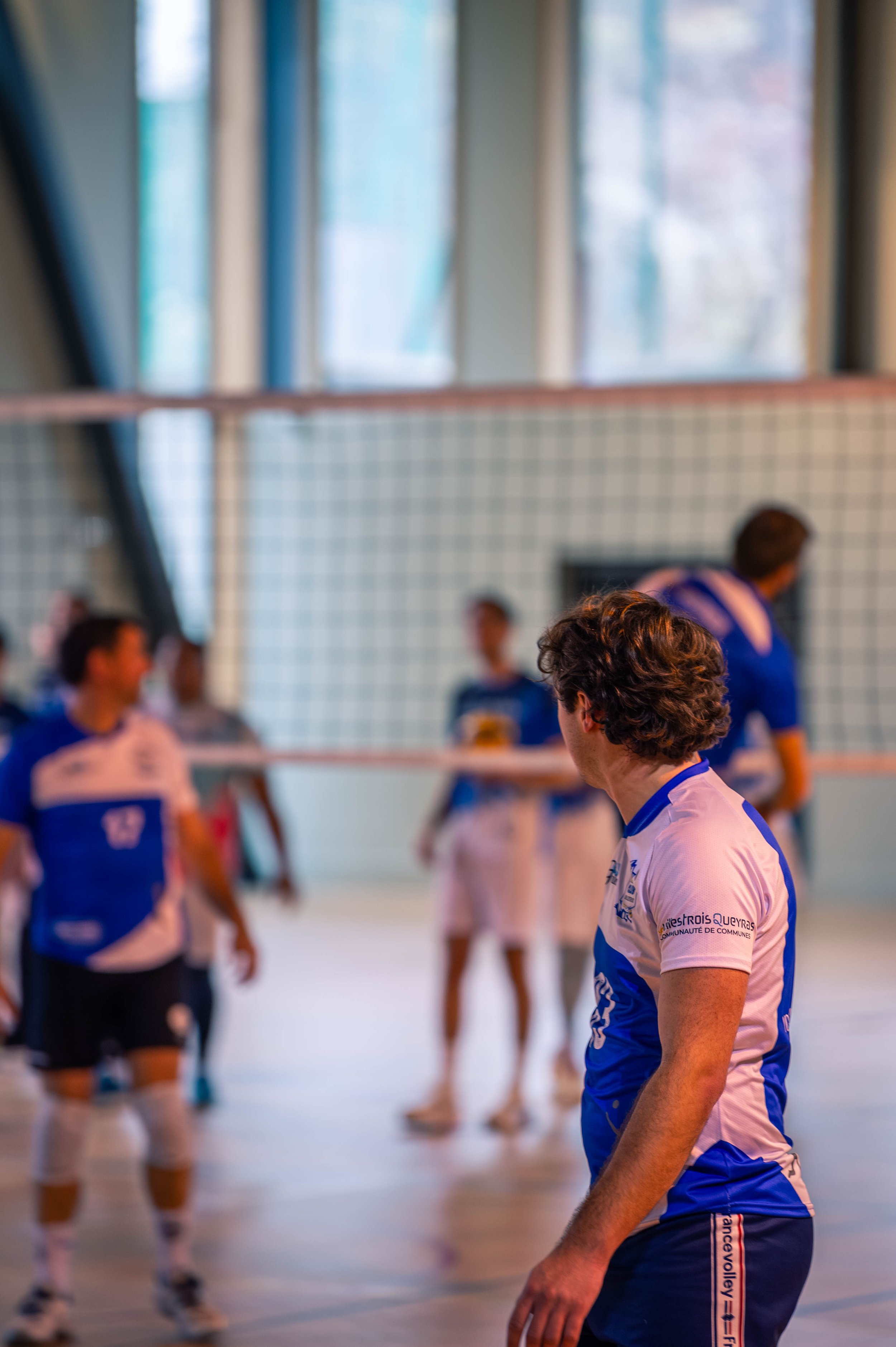 Jeu de volley-ball en gymnase avec joueurs en maillots bleus et blancs, un des joueurs au premier plan regarde vers le reste de l'équipe.