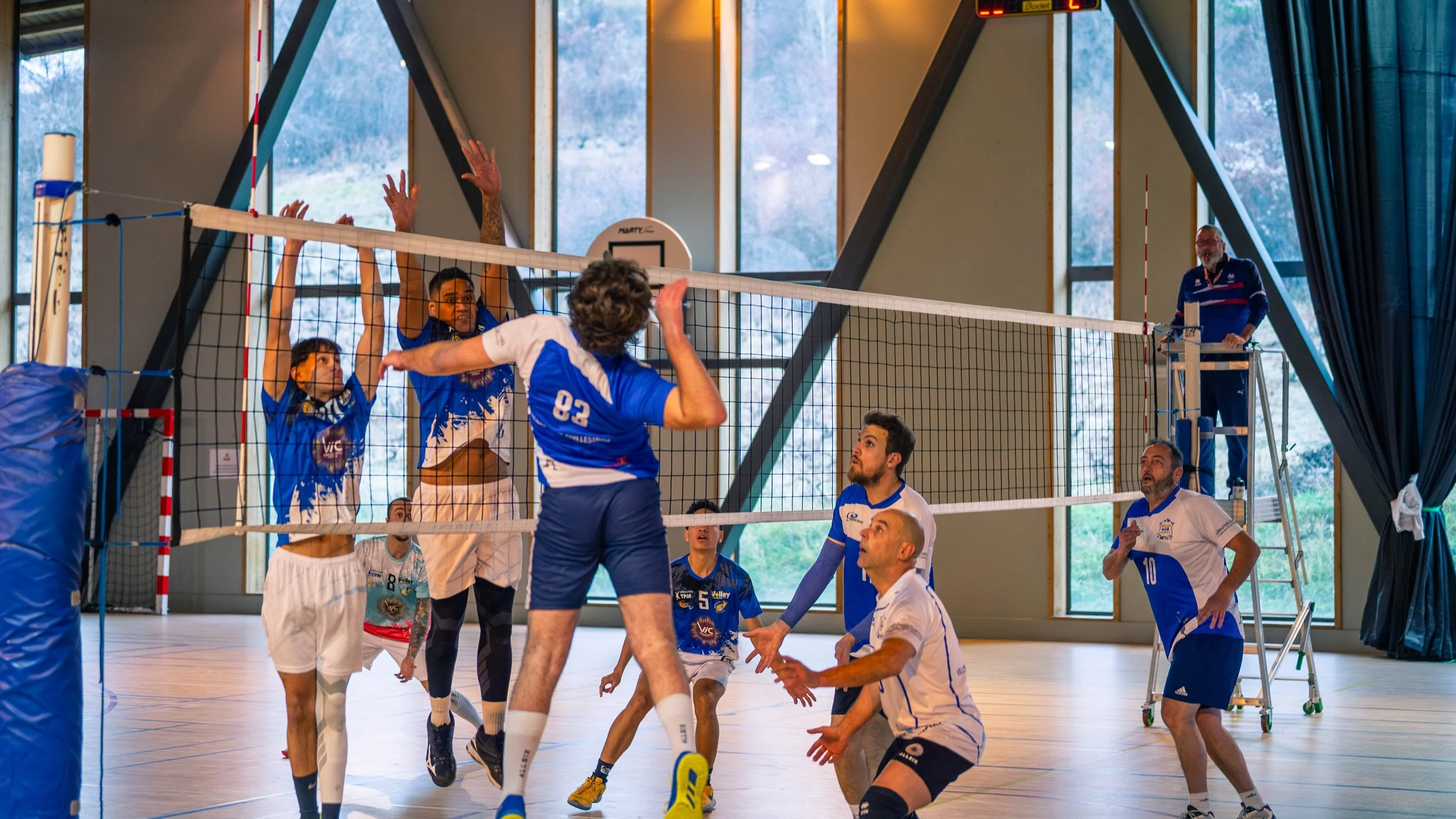 Une équipe de volleyball en train de jouer lors d'un match intérieur, avec un joueur sautant pour un spike et d'autres préparés pour une réception ou un bloc, en présence d'un arbitre sur une table.