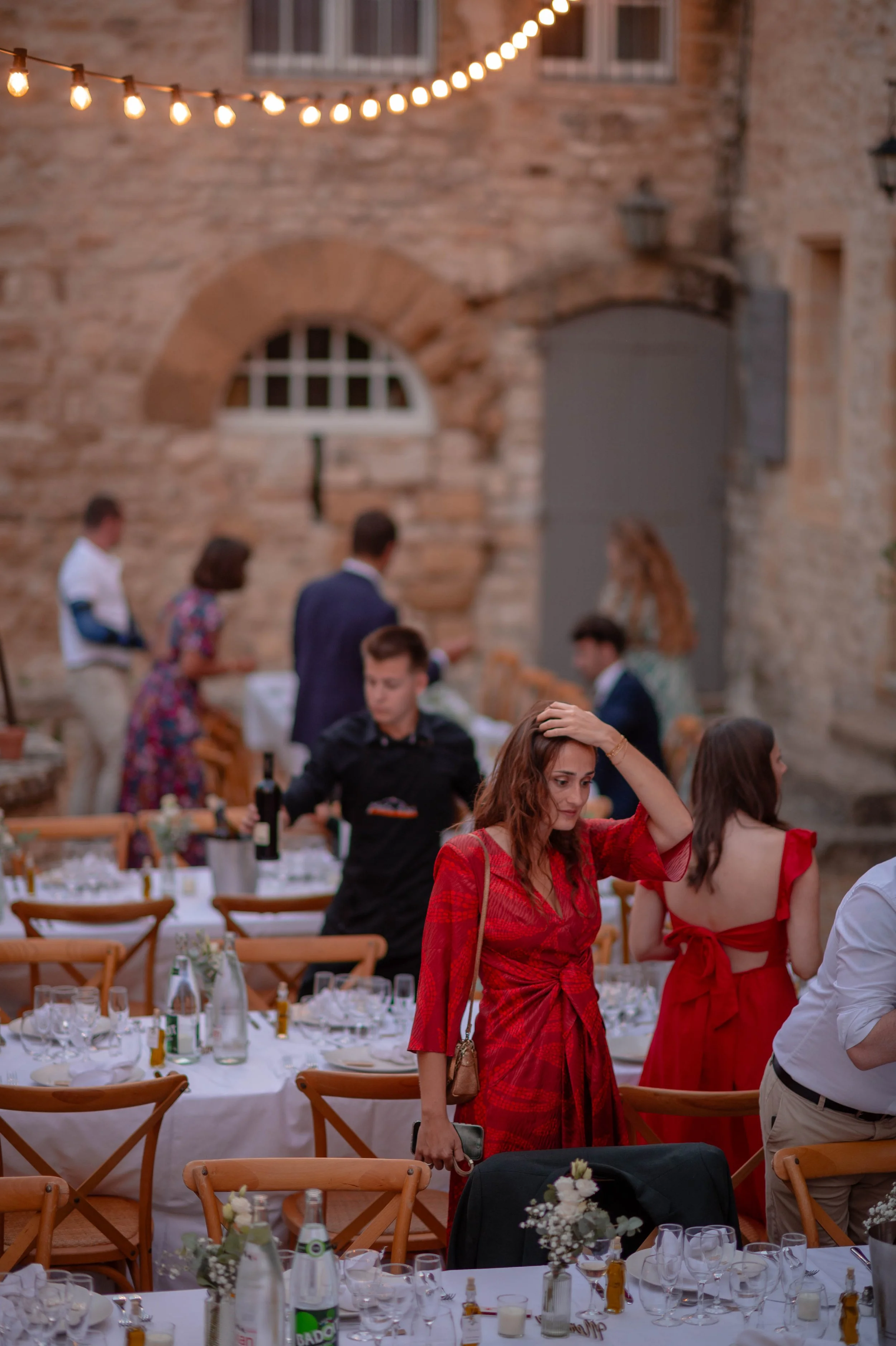 Une femme en robe rouge tient un téléphone et semble préoccupée, lors d'un repas en plein air dans une cour avec des tables dressées et des invités. La scène est éclairée par des guirlandes lumineuses.