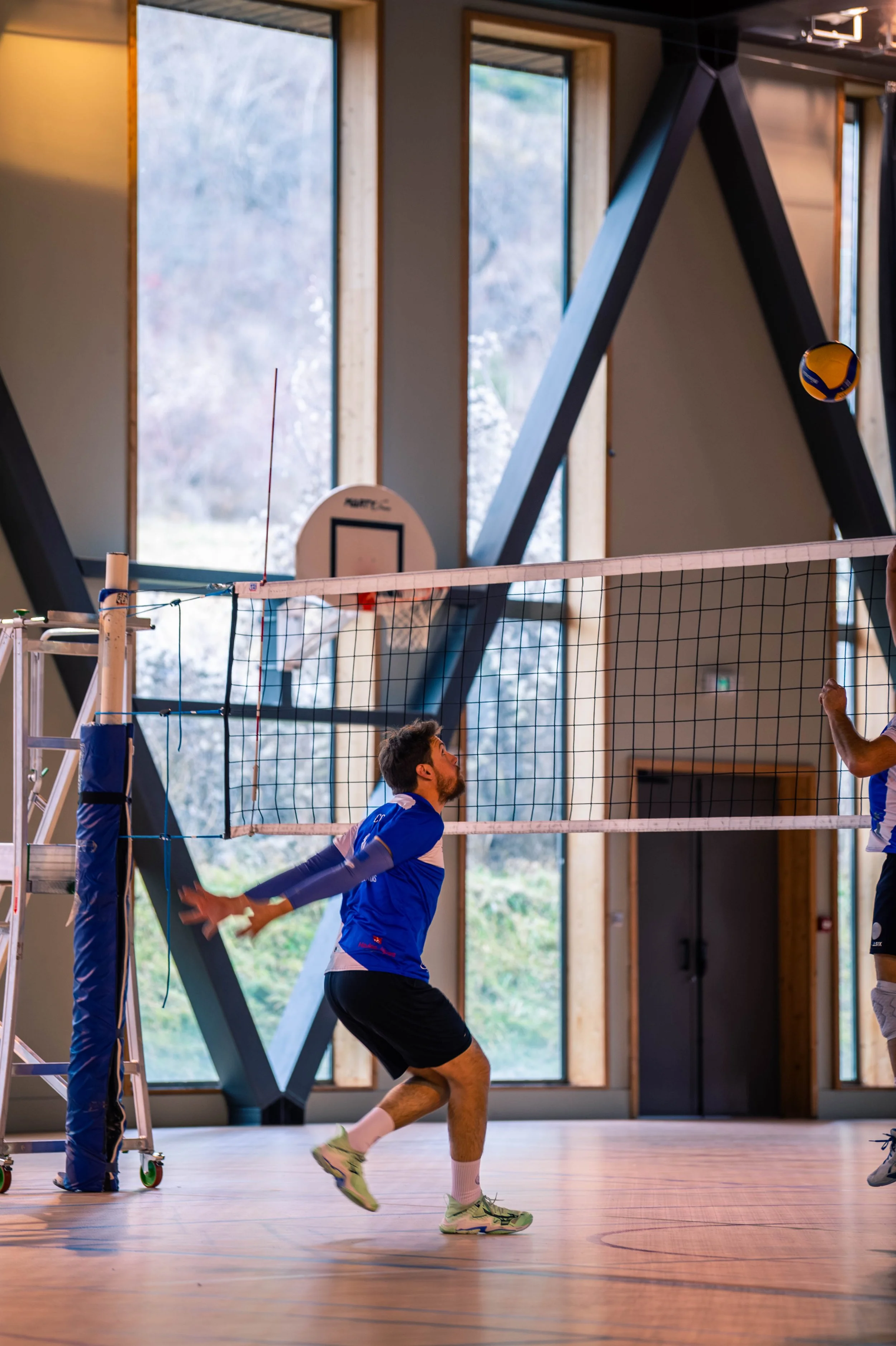 Une équipe de volleyball en entraînement dans un gymnase avec de grandes fenêtres et un panier de basket en arrière-plan.
