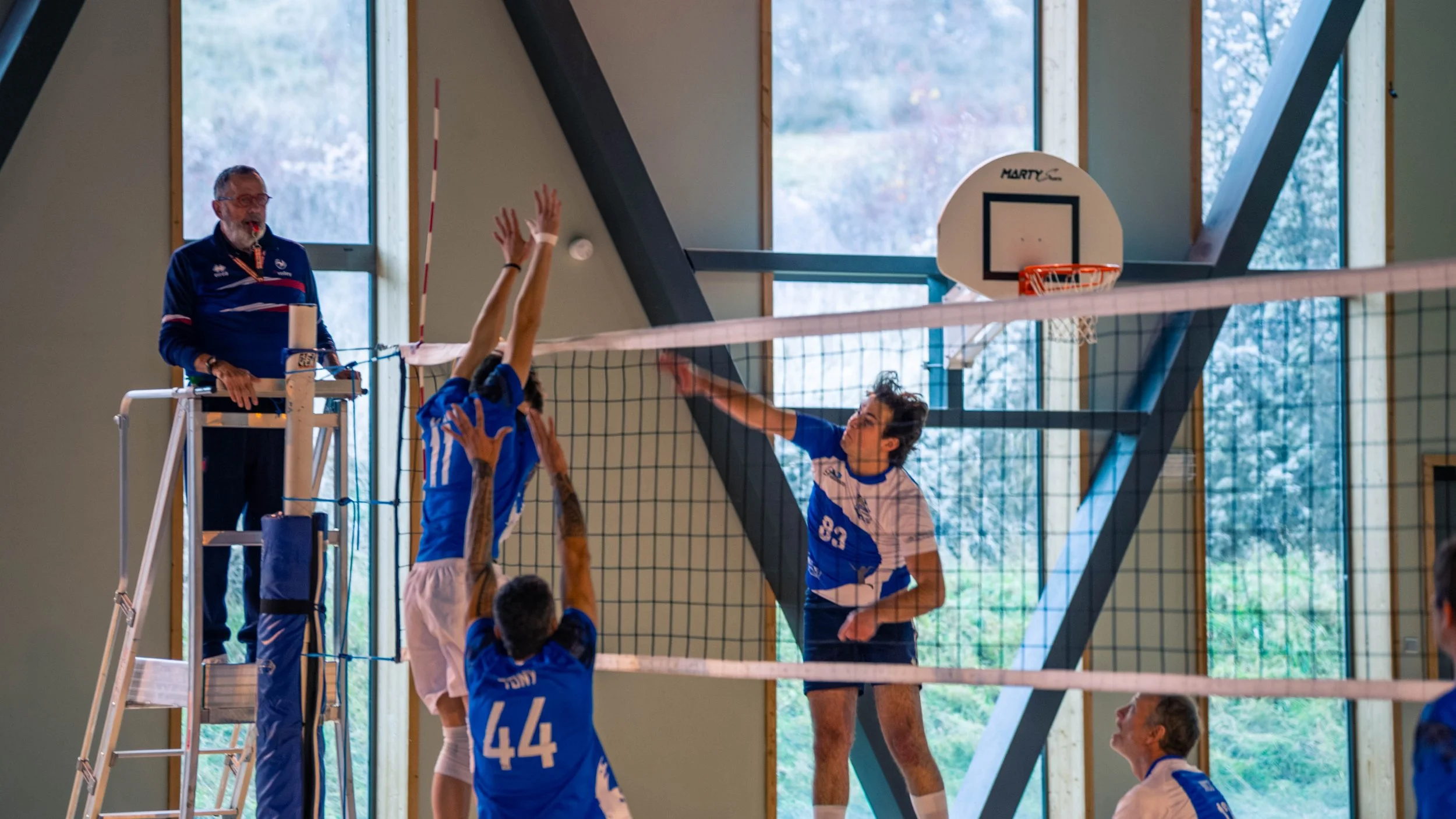 Une équipe de volleyball en action à l'intérieur d’un gymnase, avec un joueur en blanc tentant de faire passer le ballon au-dessus du filet, tandis que ses adversaires en bleu tentent de le bloquer. En arrière-plan, un arbitre en uniforme observe le 