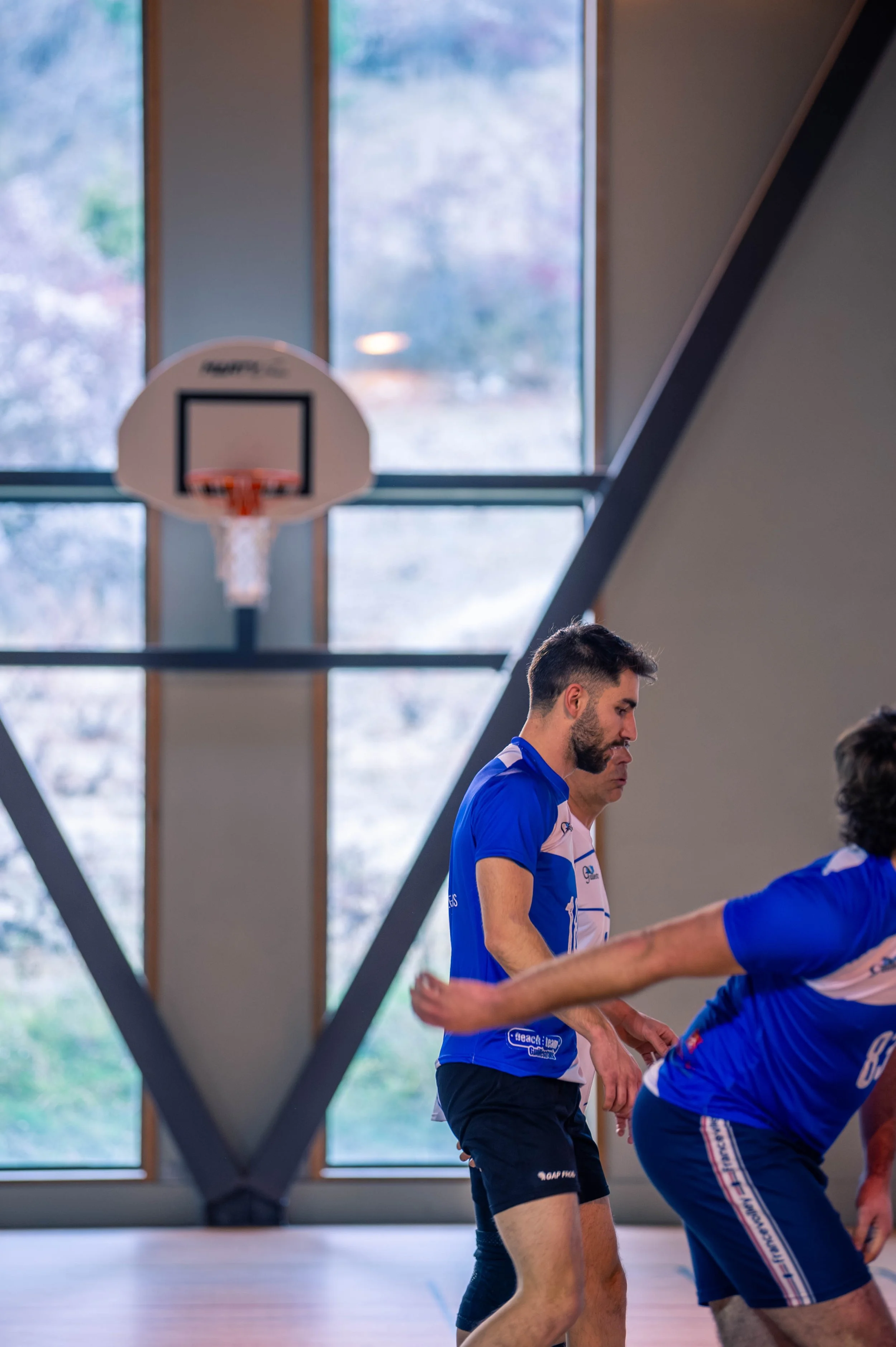 Deux hommes jouent au basketball dans une salle avec de grandes fenêtres et un tableau de basketball suspendu au mur.