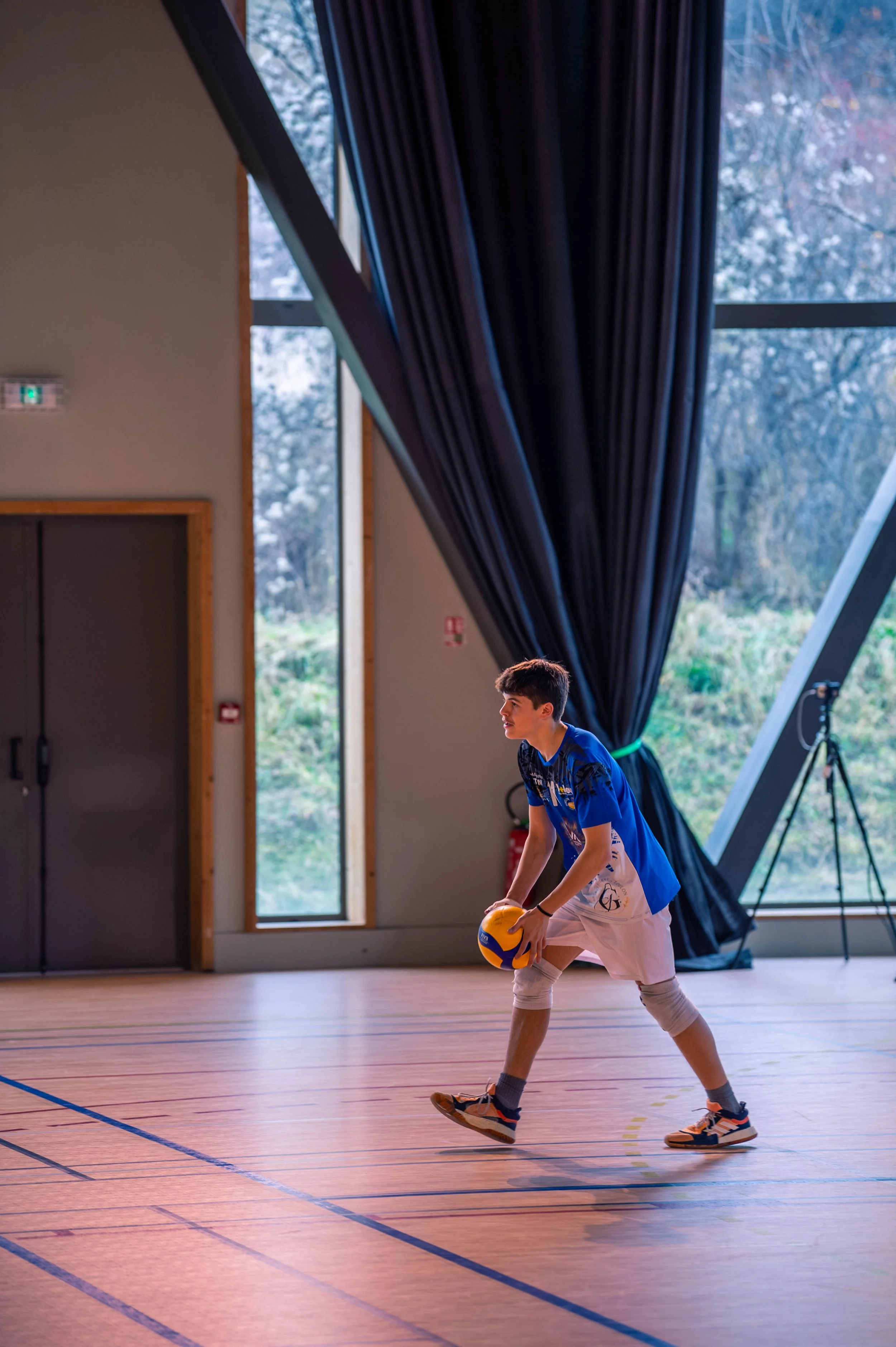Un jeune garçon joue au volley-ball dans une salle de sport intérieure, tenant un ballon de volleyball, portant un maillot bleu et un short blanc, avec de grandes fenêtres laissant entrer la lumière naturelle.
