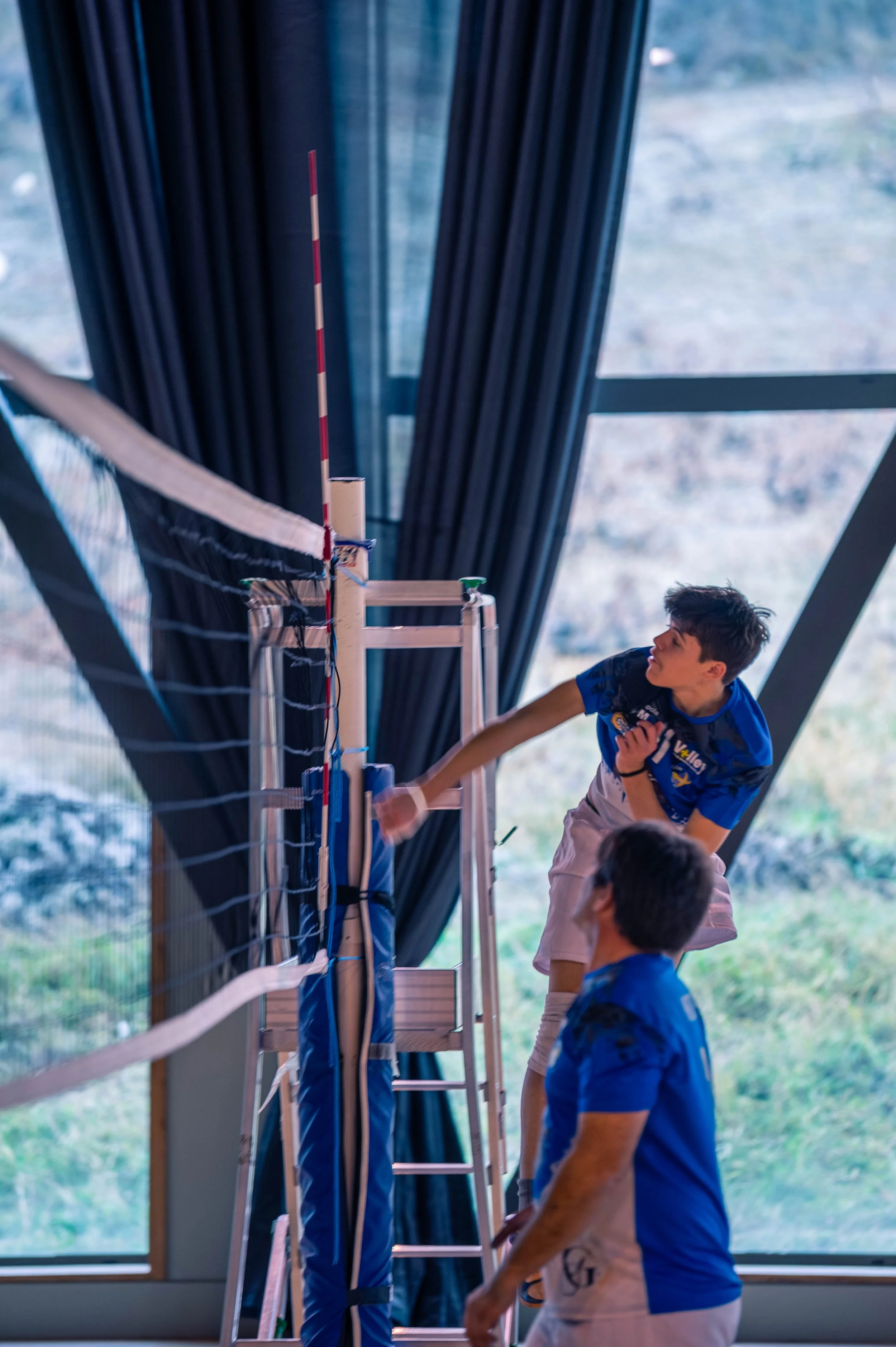 Deux jeunes garçons jouent au volleyball intérieur, l'un d'eux saute pour toucher le ballon près du filet, dans une salle avec de grandes fenêtres et des rideaux noirs.