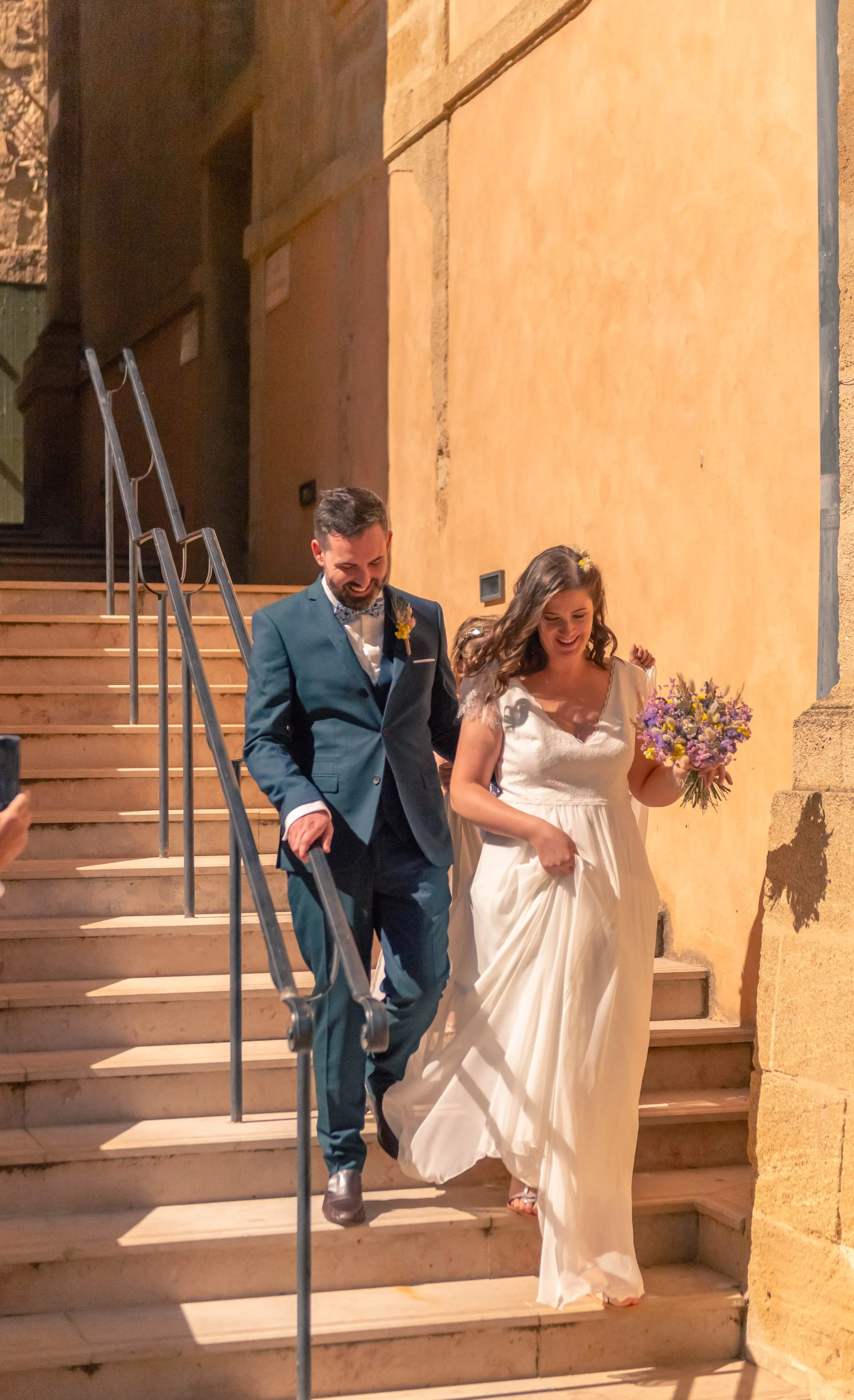 Un couple de mariés descend des escaliers en pierre à l'extérieur en soirée, la mariée tient un bouquet de fleurs violettes et porte une robe blanche, l'homme porte un costume bleu foncé.