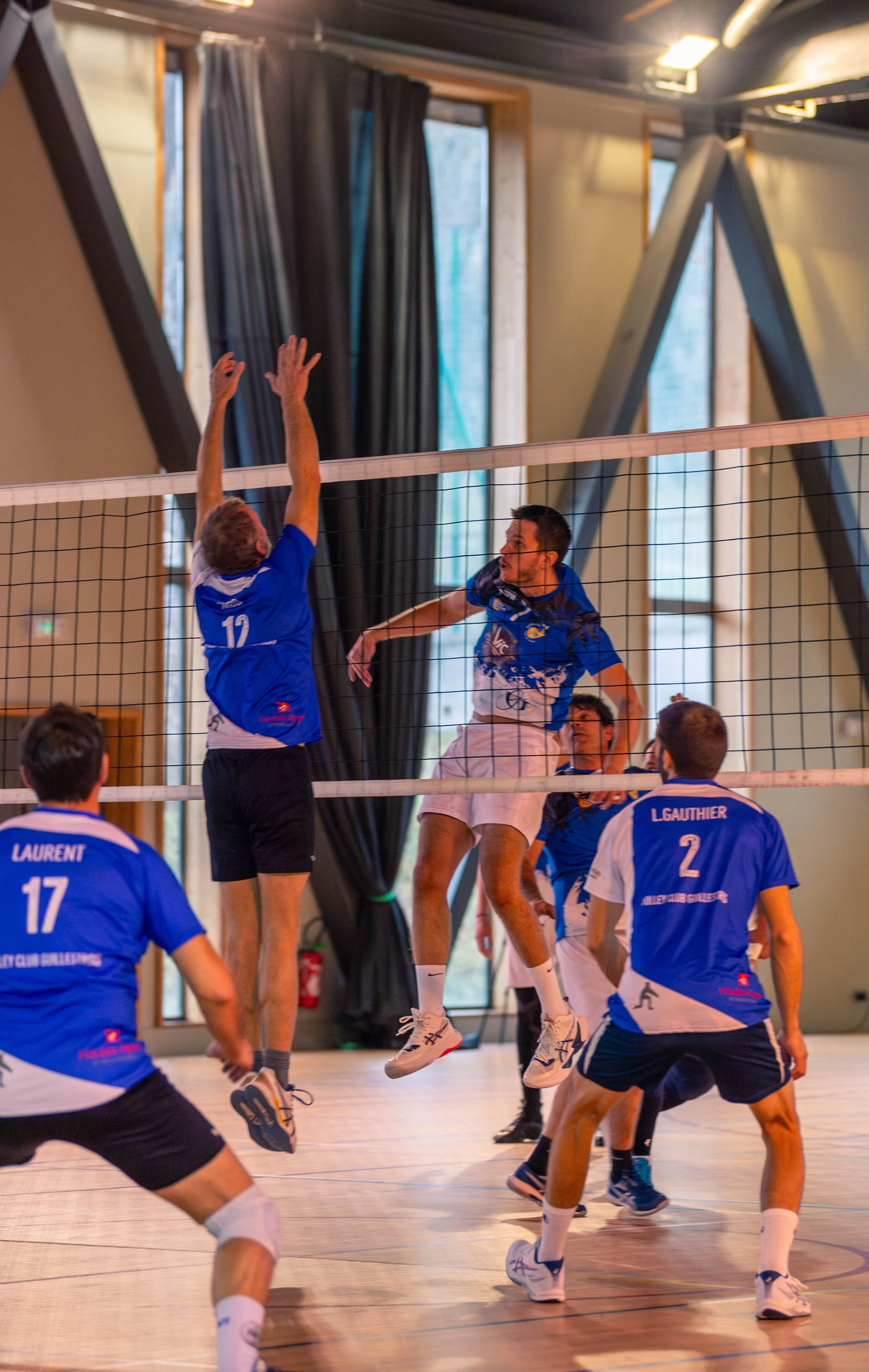 Des joueurs de volley-ball en action lors d'un match intérieur; un joueur est en saut pour attaquer le ballon tandis qu'un autre de l'équipe adverse tente de le bloquer.
