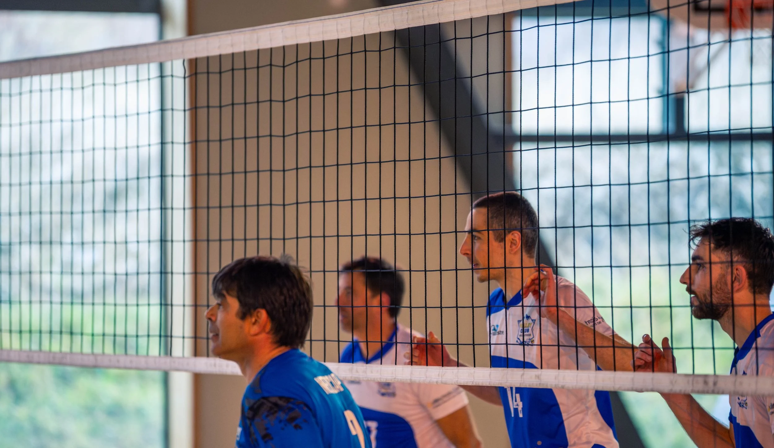 Joueurs de volleyball en préparation dans une salle intérieure, vus à travers un filet.