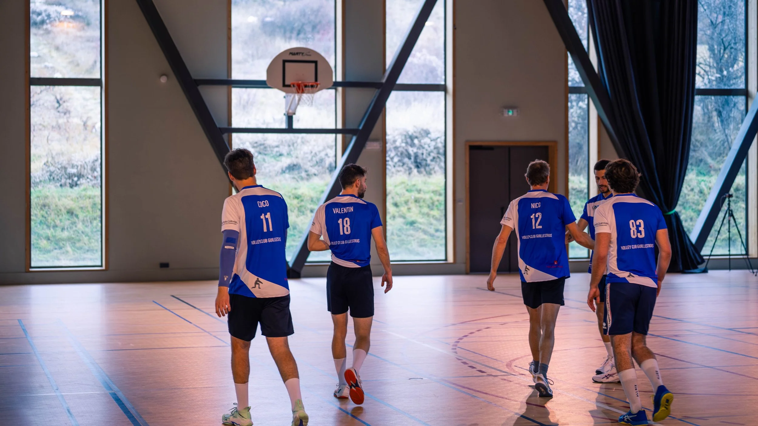 Cinq joueurs de volley-ball dans une salle d'entraînement, portant des maillots bleus avec leurs noms et numéros, discutant près du panier de basketball, avec de grandes fenêtres laissant entrer la lumière naturelle.