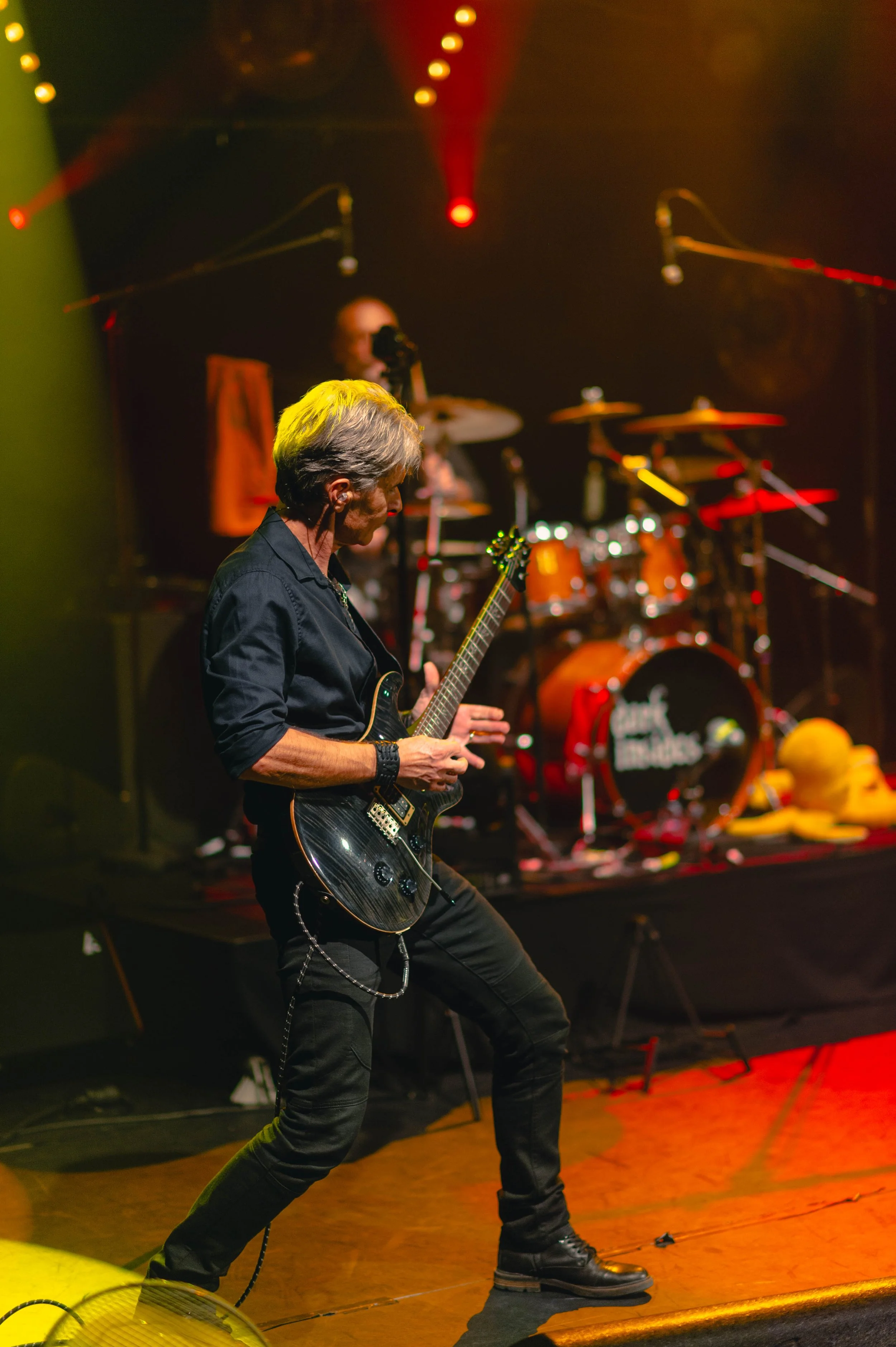 Musicien joué de la guitare électrique sur scène lors d'un concert, avec un batteur en arrière-plan, sous des lumières colorées.