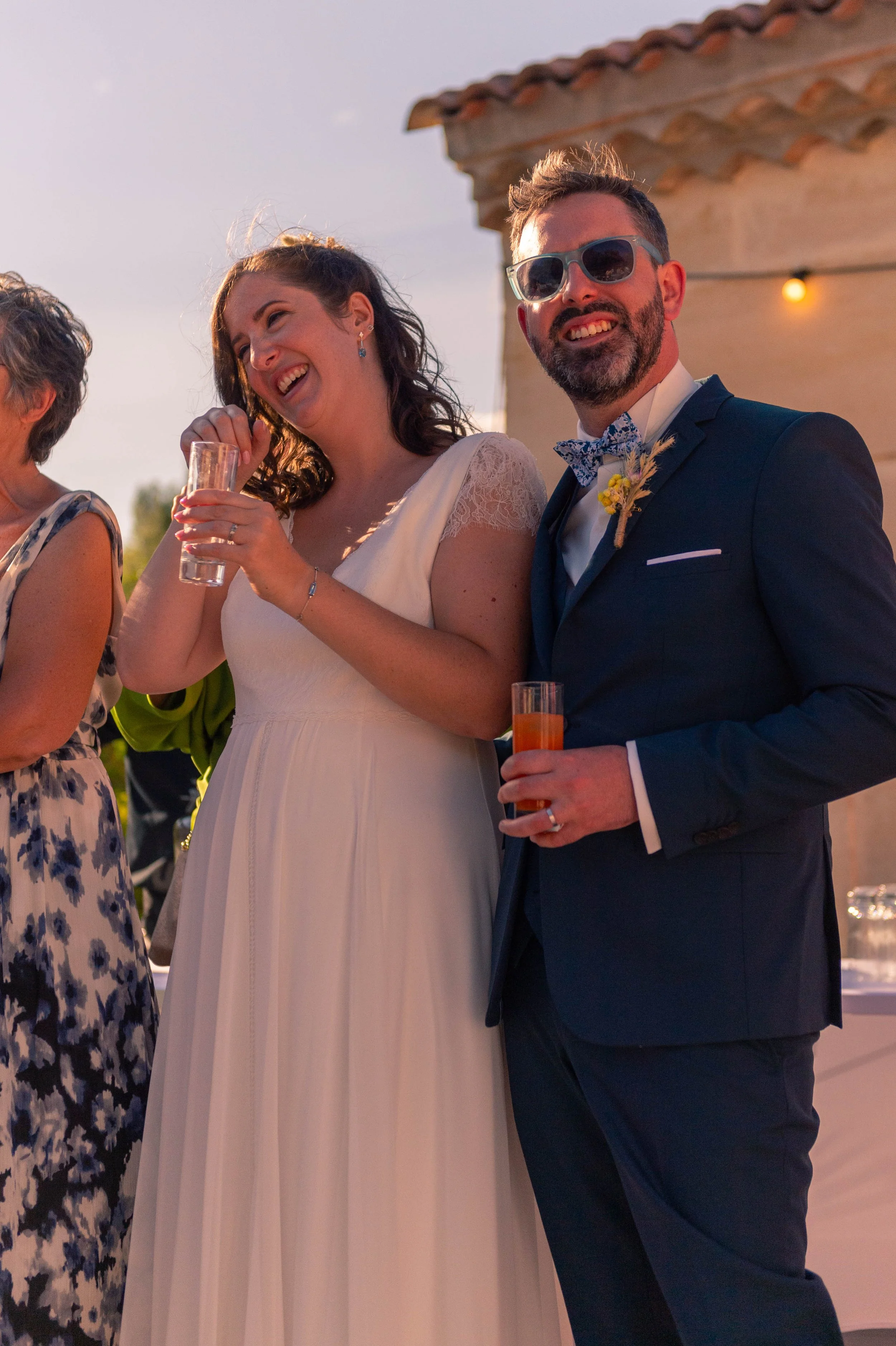 Un couple lors d'un mariage en soirée, homme en costume bleu avec lunettes de soleil, femme en robe blanche, tenant un verre, souriant.
