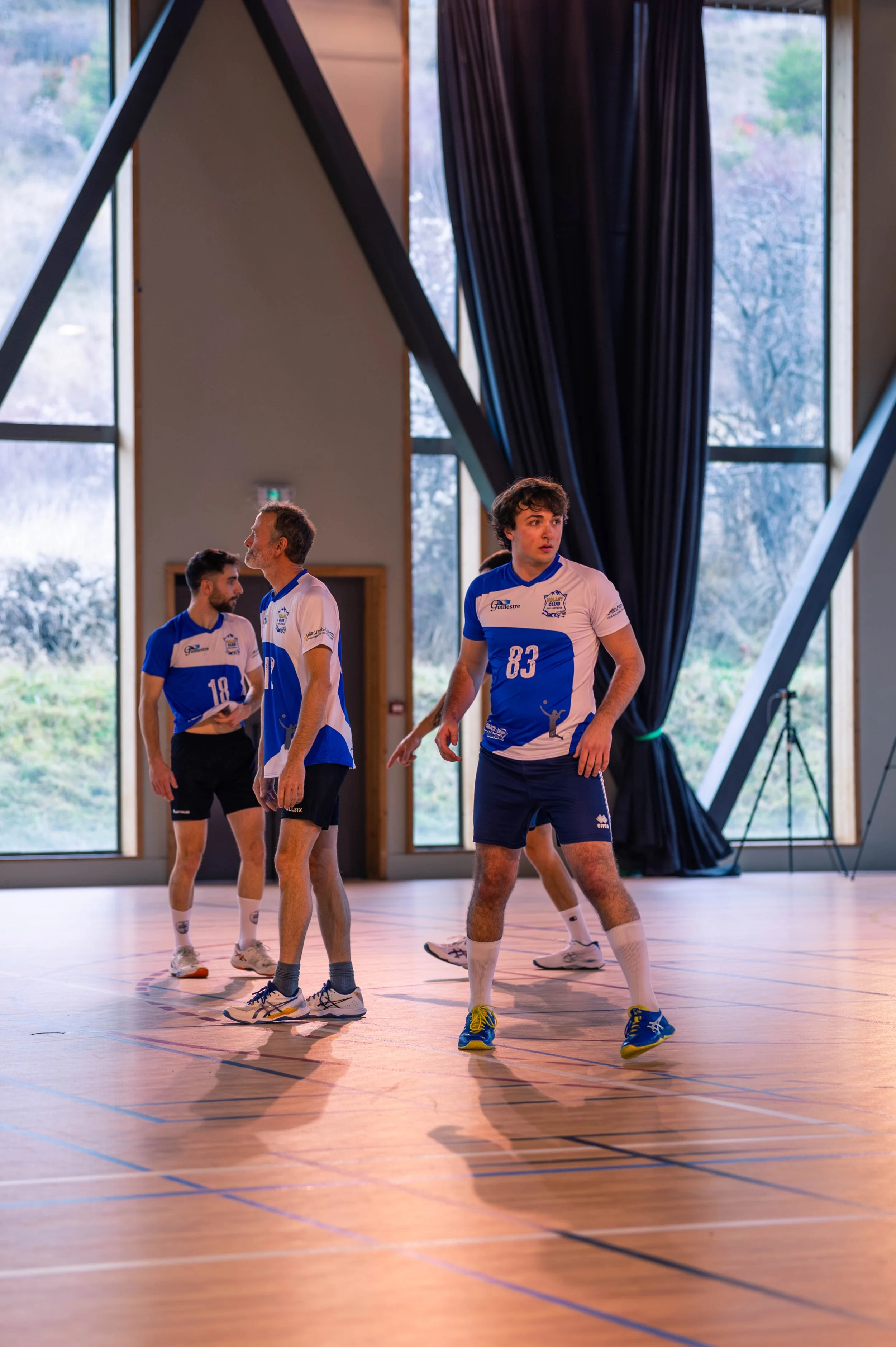 Groupe de jeunes athlètes en entraînement dans une salle de sport intérieure, vêtus de maillots bleus et blancs, sur un parquet de gym, avec de grandes fenêtres laissant entrer la lumière naturelle.