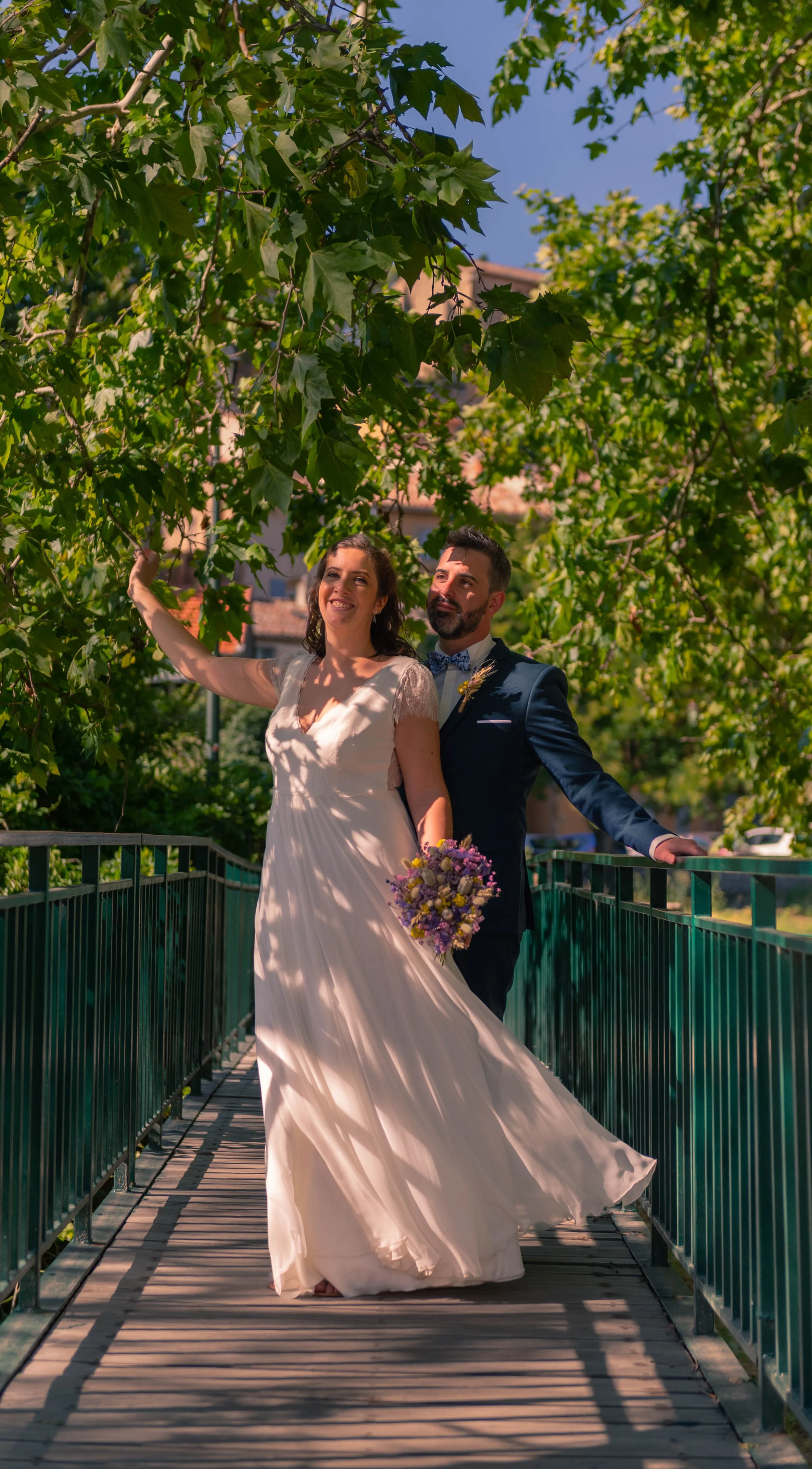 Un couple de mariés posant sur un pont en extérieur, entouré d'arbres, avec la mariée en robe blanche et le marié en costume bleu, la mariée tenant un bouquet de fleurs.