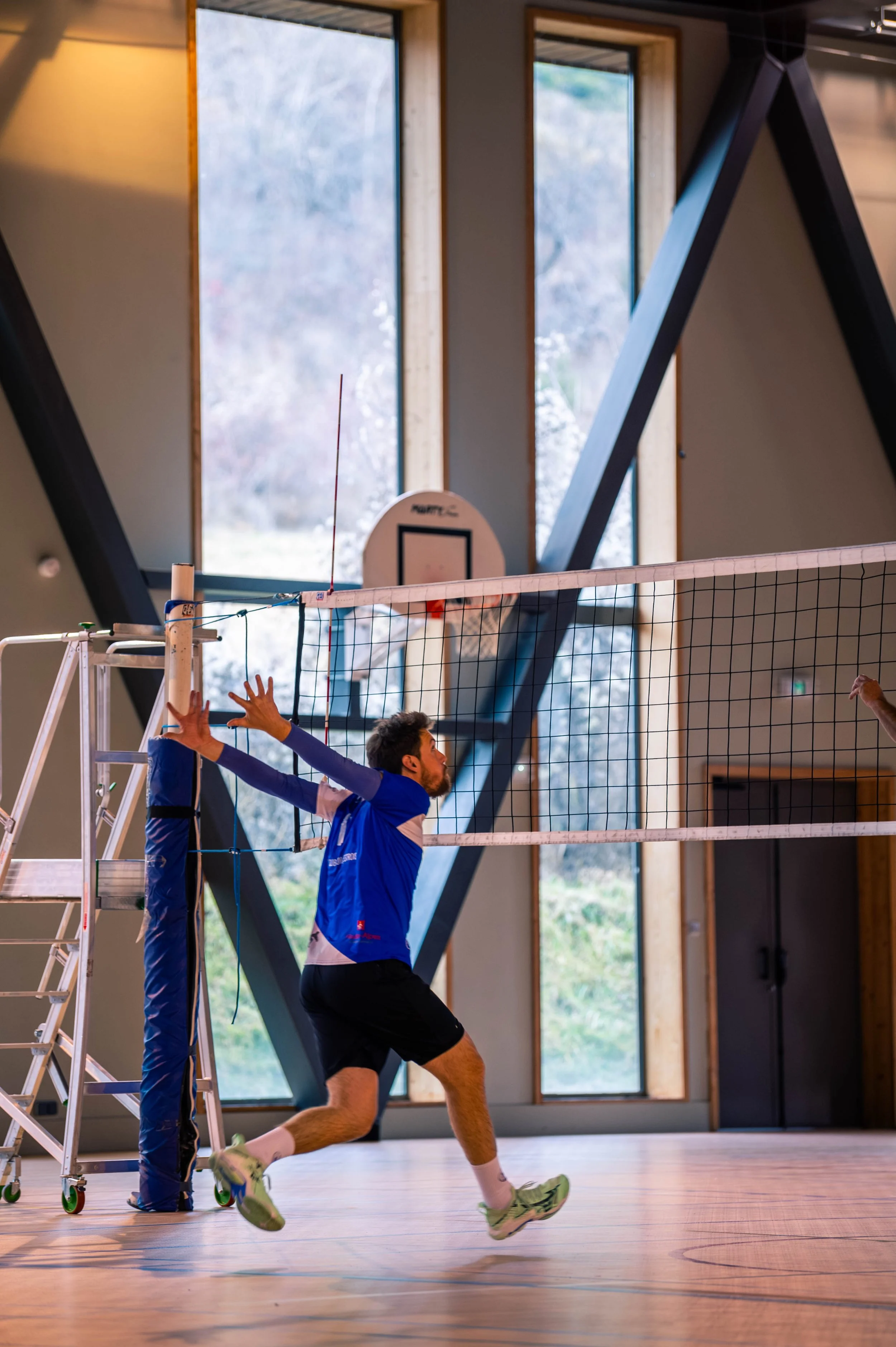 Un homme joue au volleyball en intérieur, prêt à frapper le ballon, dans un gymnase avec de grandes fenêtres et un panneau de basketball en fond.