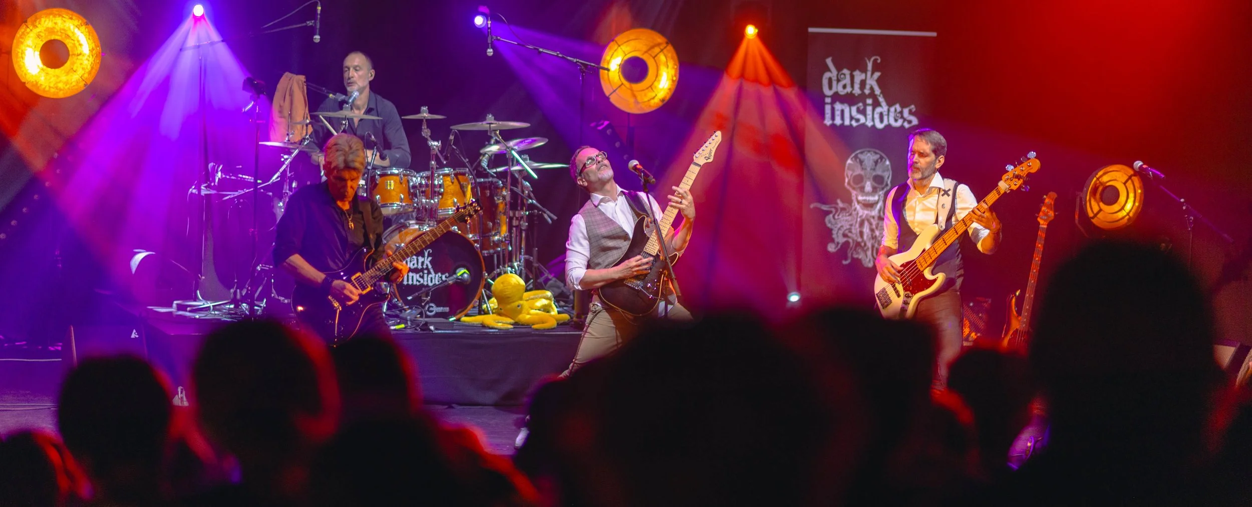 Un groupe de musiciens jouant sur scène lors d'un concert, avec des lumières colorées et un fond affichant le logo 'Dark Inside'.