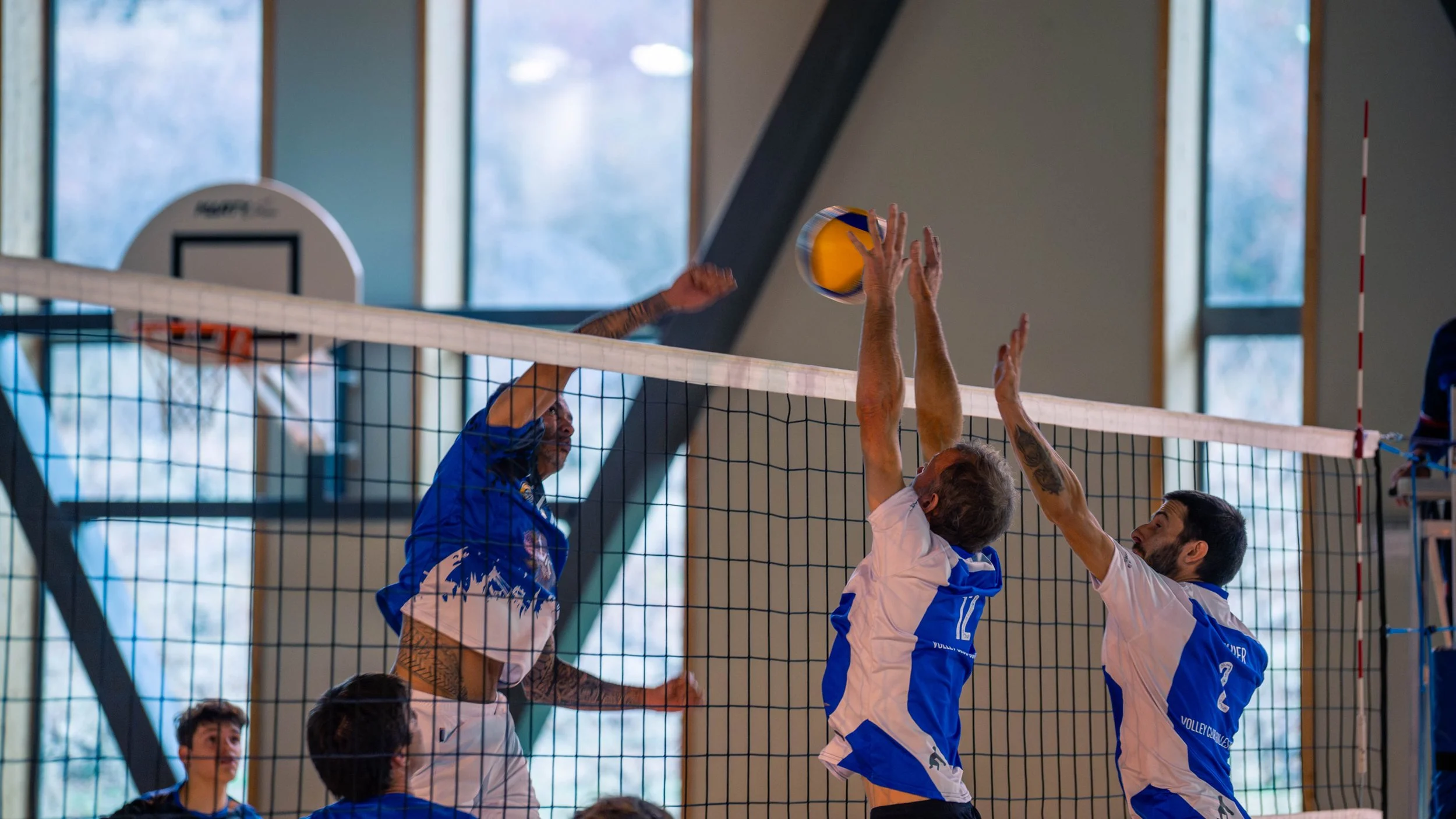Joueur en volley-ball effectuant un smash au-dessus du filet face à deux défenseurs qui tentent de bloquer le ballon, dans une salle de sport.