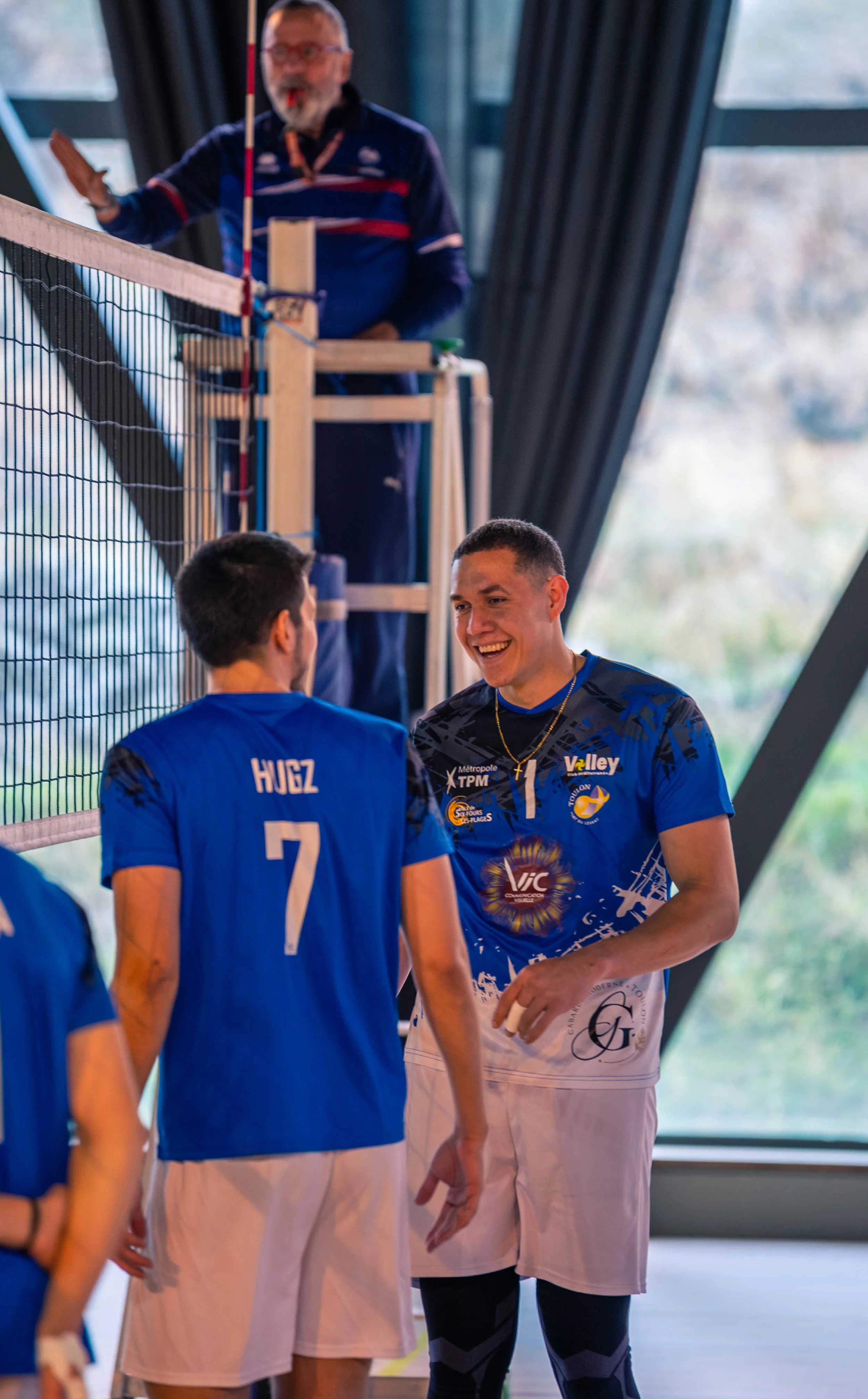 Joueurs de volleyball en tenue bleue, souriants, en discussion lors d'un match ou entraînement, avec un arbitre en arrière-plan dans une salle intérieure.