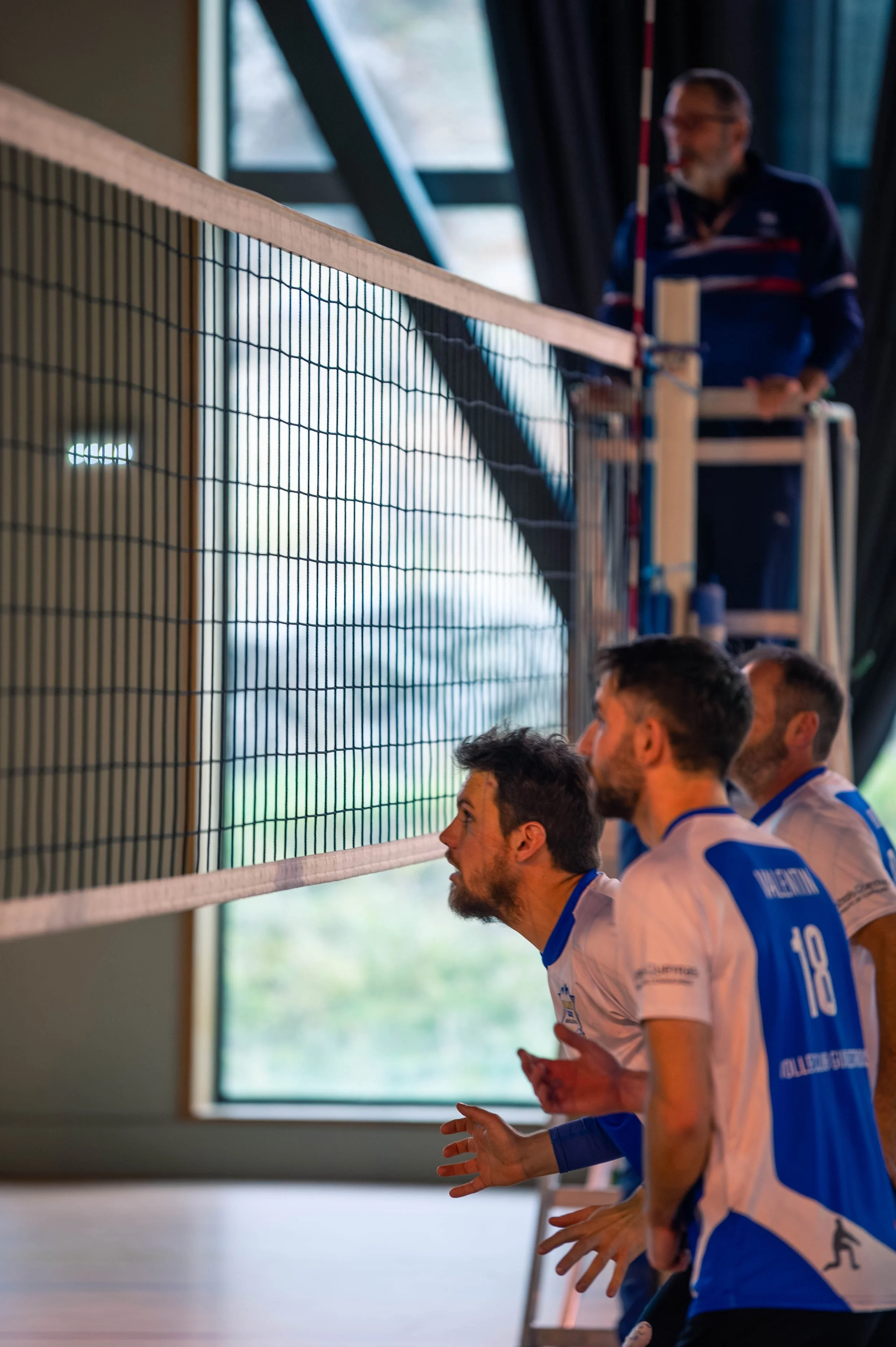 Trois joueurs de volley-ball en position d'attente devant un filet, en train de recevoir une balle dans un gymnase, avec un entraîneur sur une plateforme de remontée en arrière-plan.