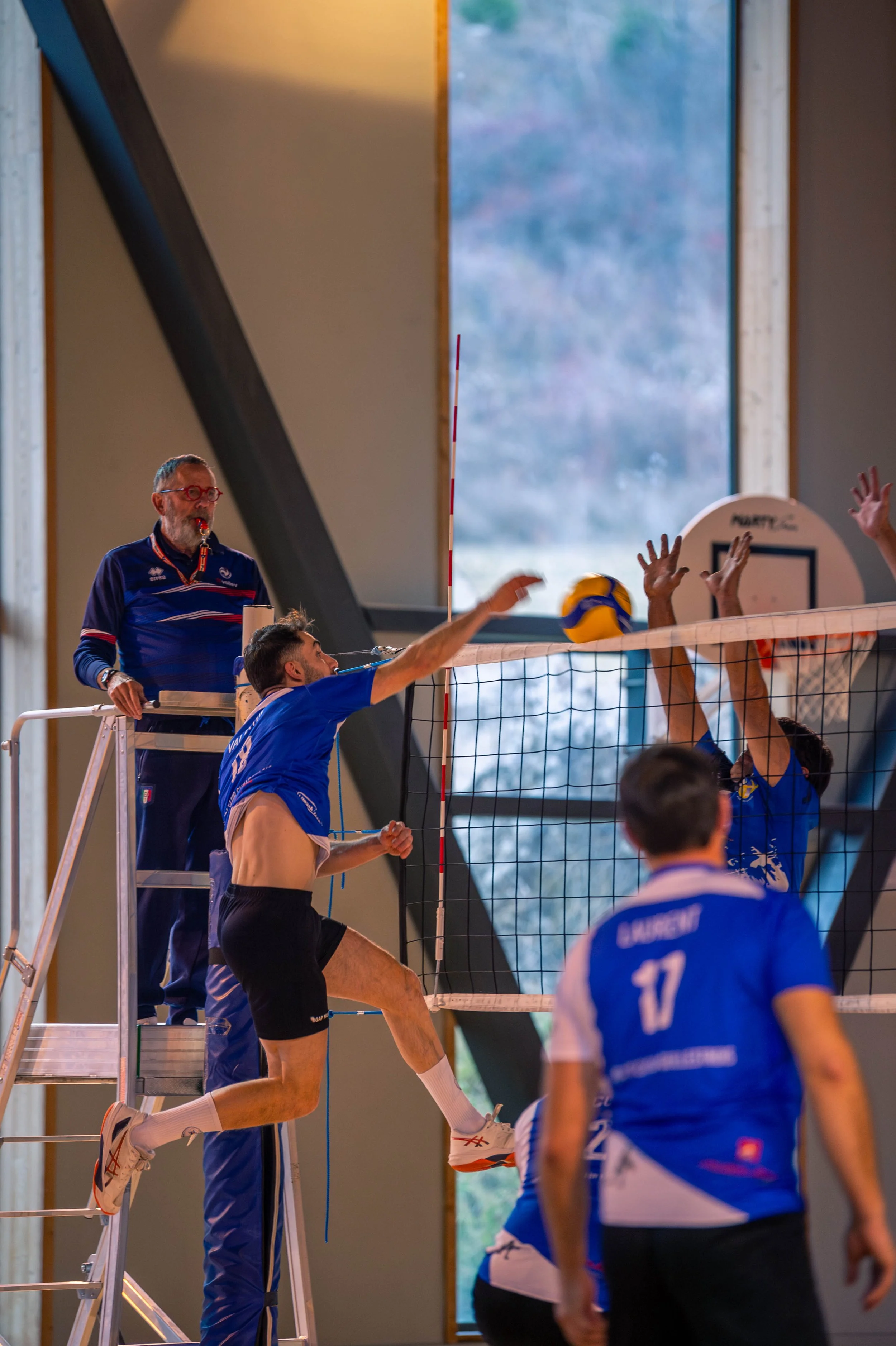 Match de volleyball en intérieur avec joueurs en action, un arbitre sur une plateforme, et un mur avec une grande fenêtre en arrière-plan.